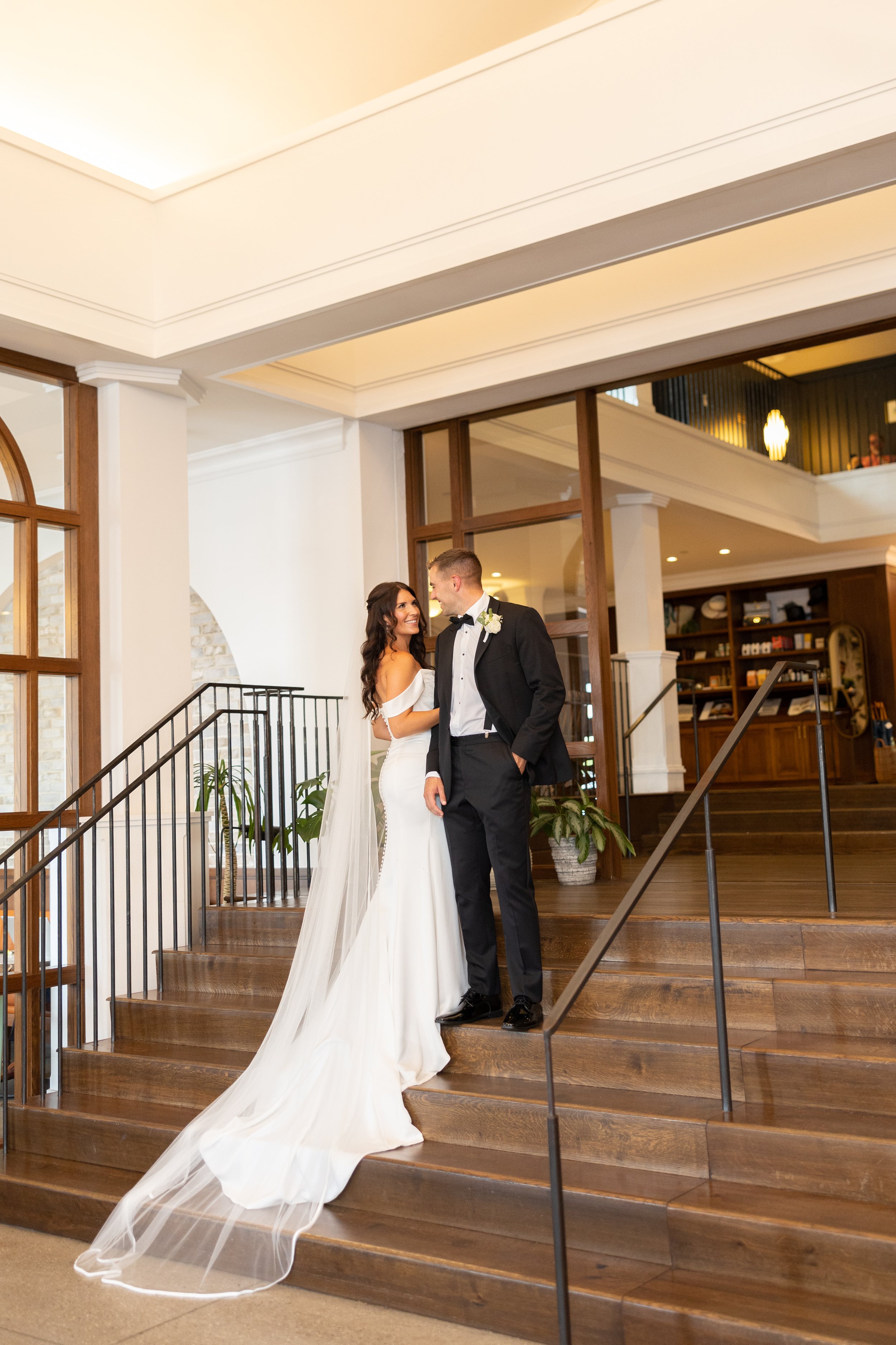 A bride in a white wedding gown and a groom in a black tuxedo standing on wooden stairs inside a well-lit venue, looking at each other and smiling.