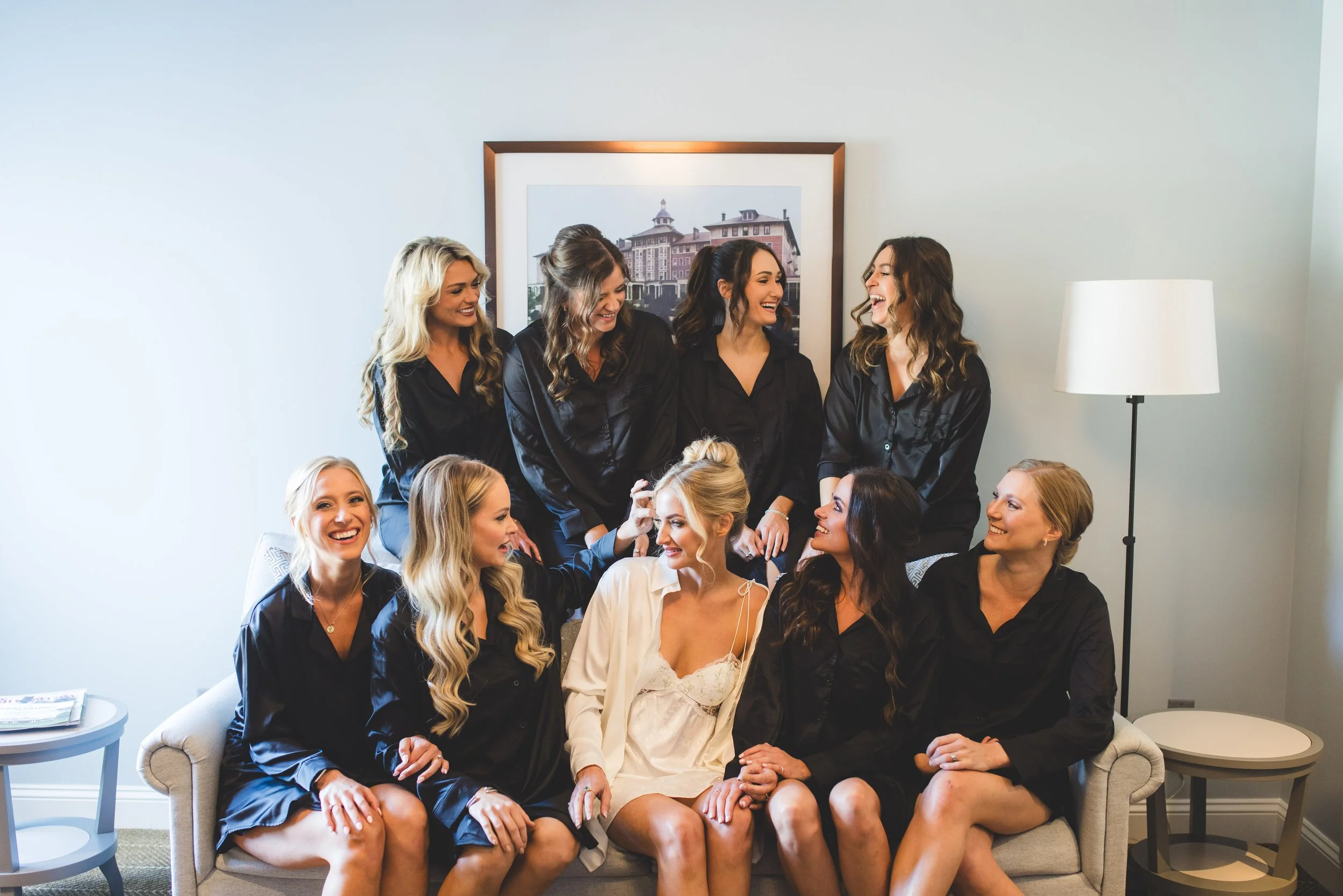Group of women in black pajamas, with one bride in a white gown, sitting and standing around on a beige sofa in a room with framed artwork on the wall, laughing and smiling, preparing for a wedding or special event.