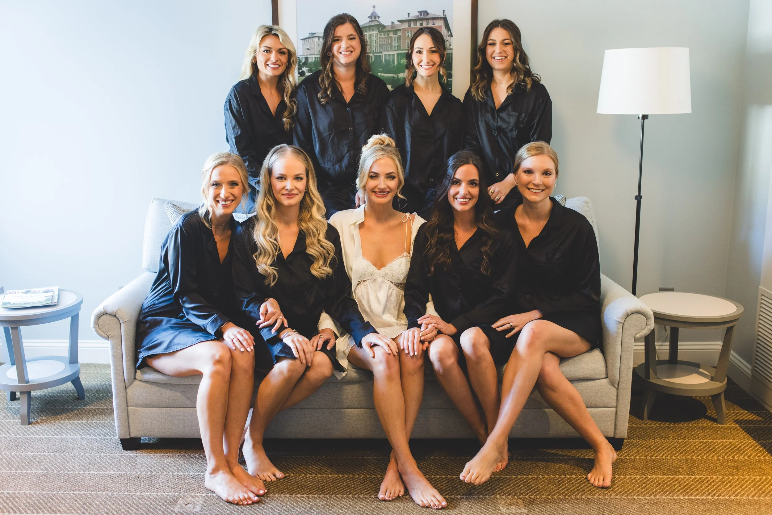 A group of ten women, mostly in matching black pajamas, sitting on a beige sofa and standing behind it, smiling in a well-lit room with a framed picture of houses on the wall and a white lamp.