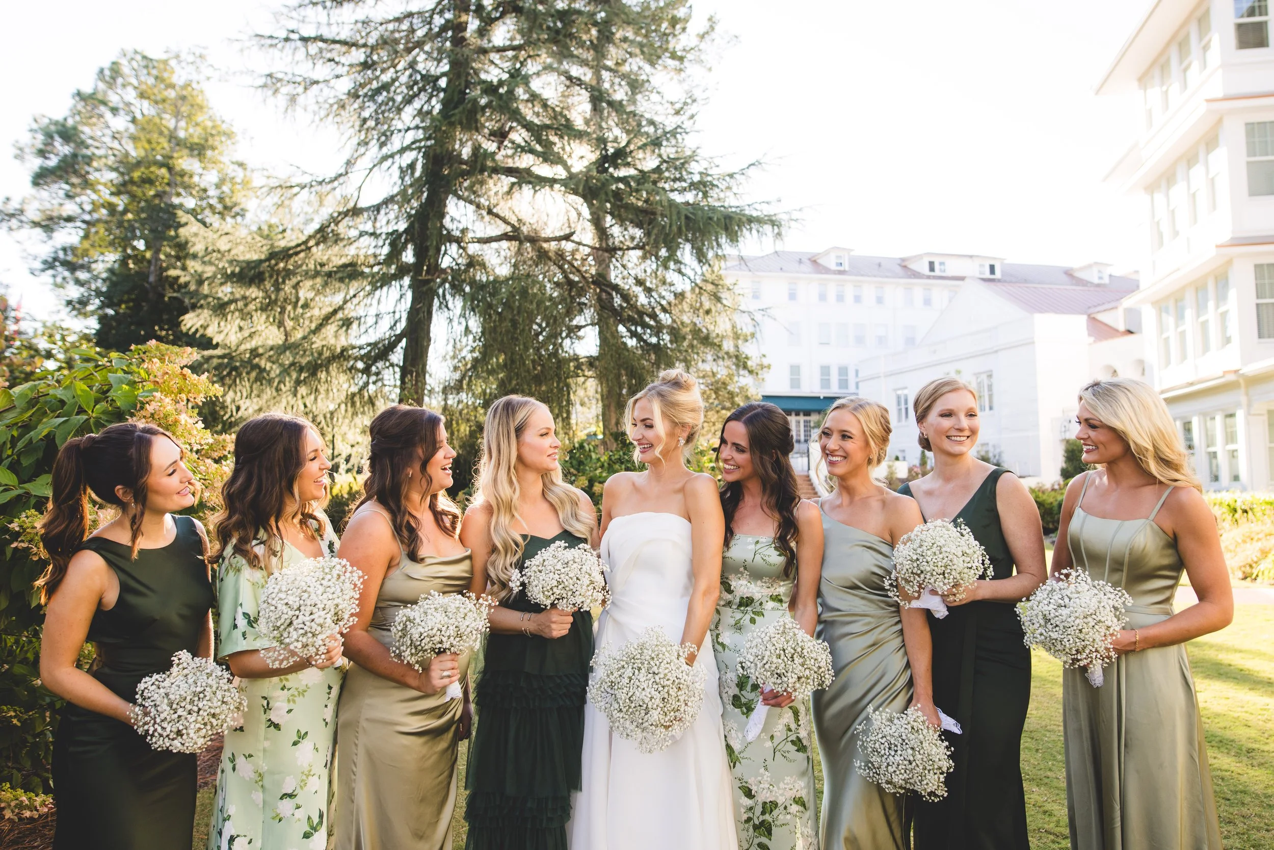 A bride in a white wedding dress standing outside with her bridesmaids holding bouquets of white baby's breath flowers.
