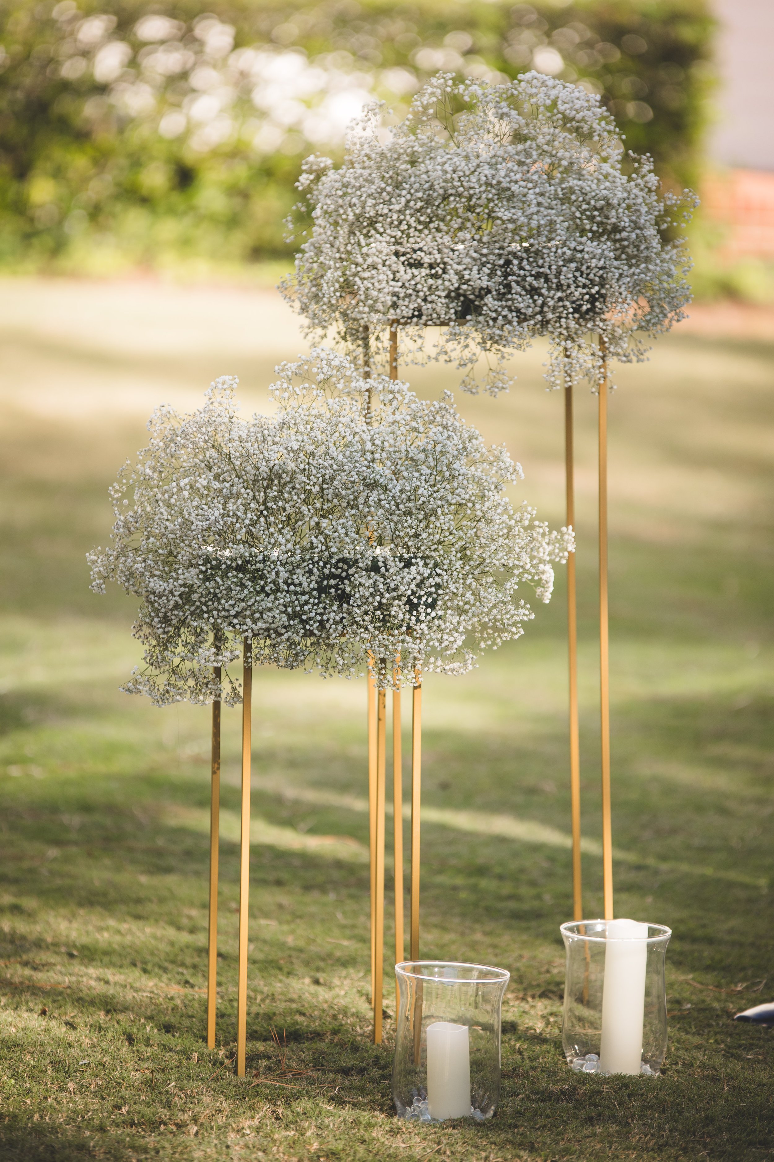 Two tall stands with small white floral arrangements, and two glass hurricane lanterns with white candles, placed outdoors on grass.