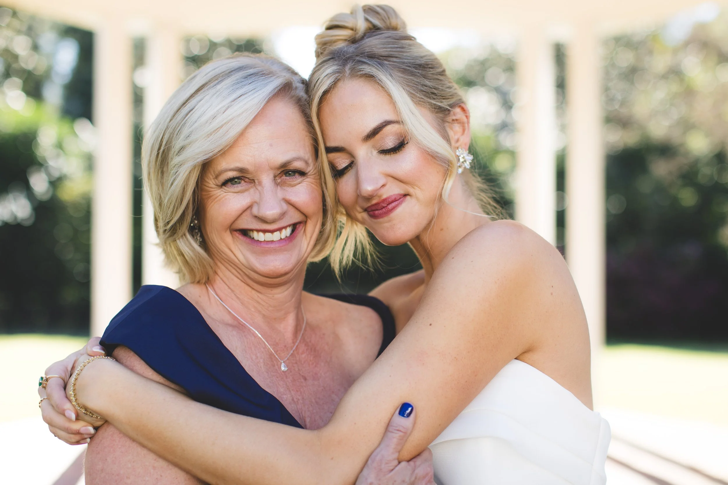 A smiling older woman and a younger woman in a white dress hug outdoors with a background of trees and white columns.