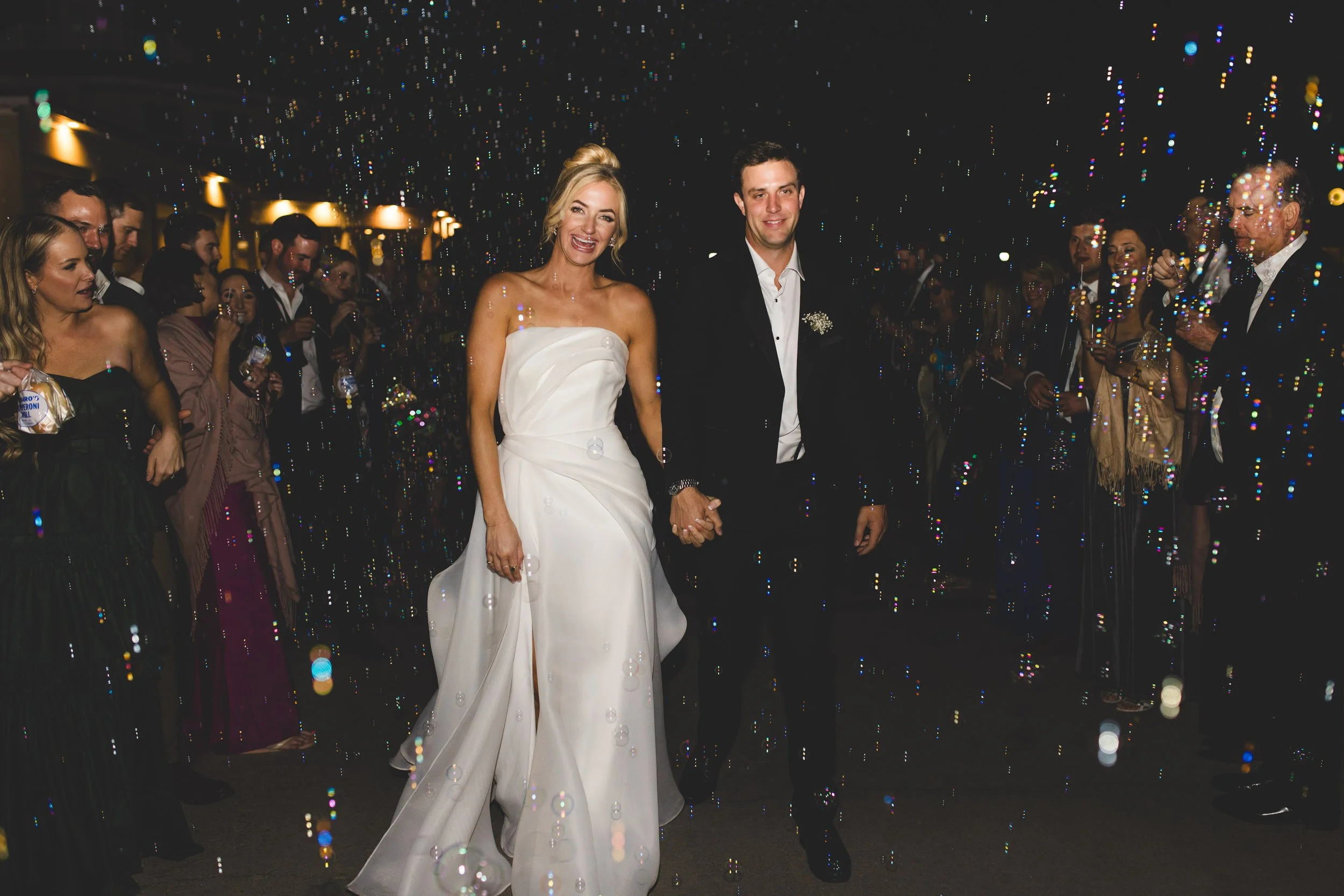 A bride and groom holding hands and smiling as they walk through a crowd of wedding guests in a celebration with confetti and bubbles.