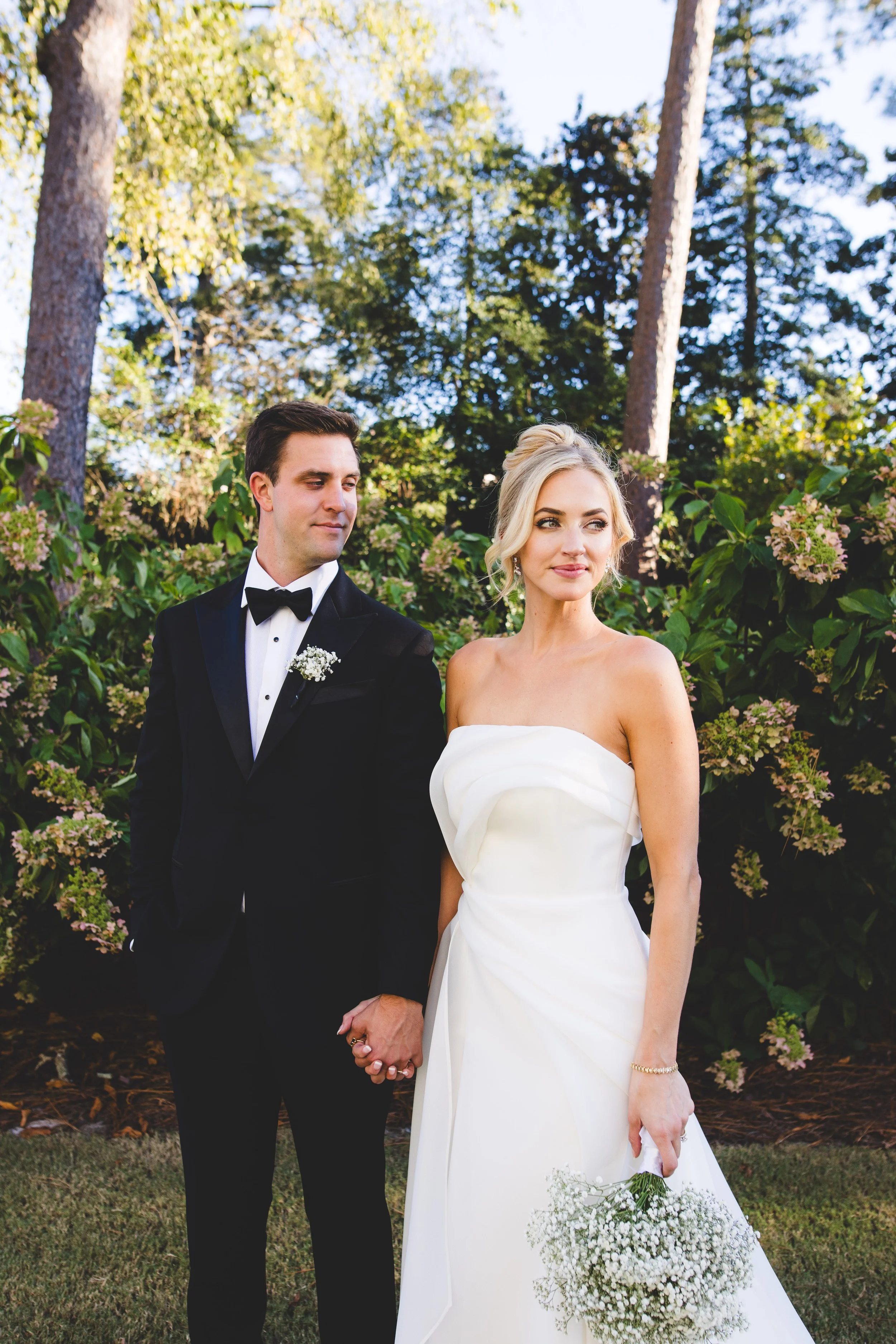 A bride and groom standing hand in hand outdoors, with trees and bushes in the background during a wedding.