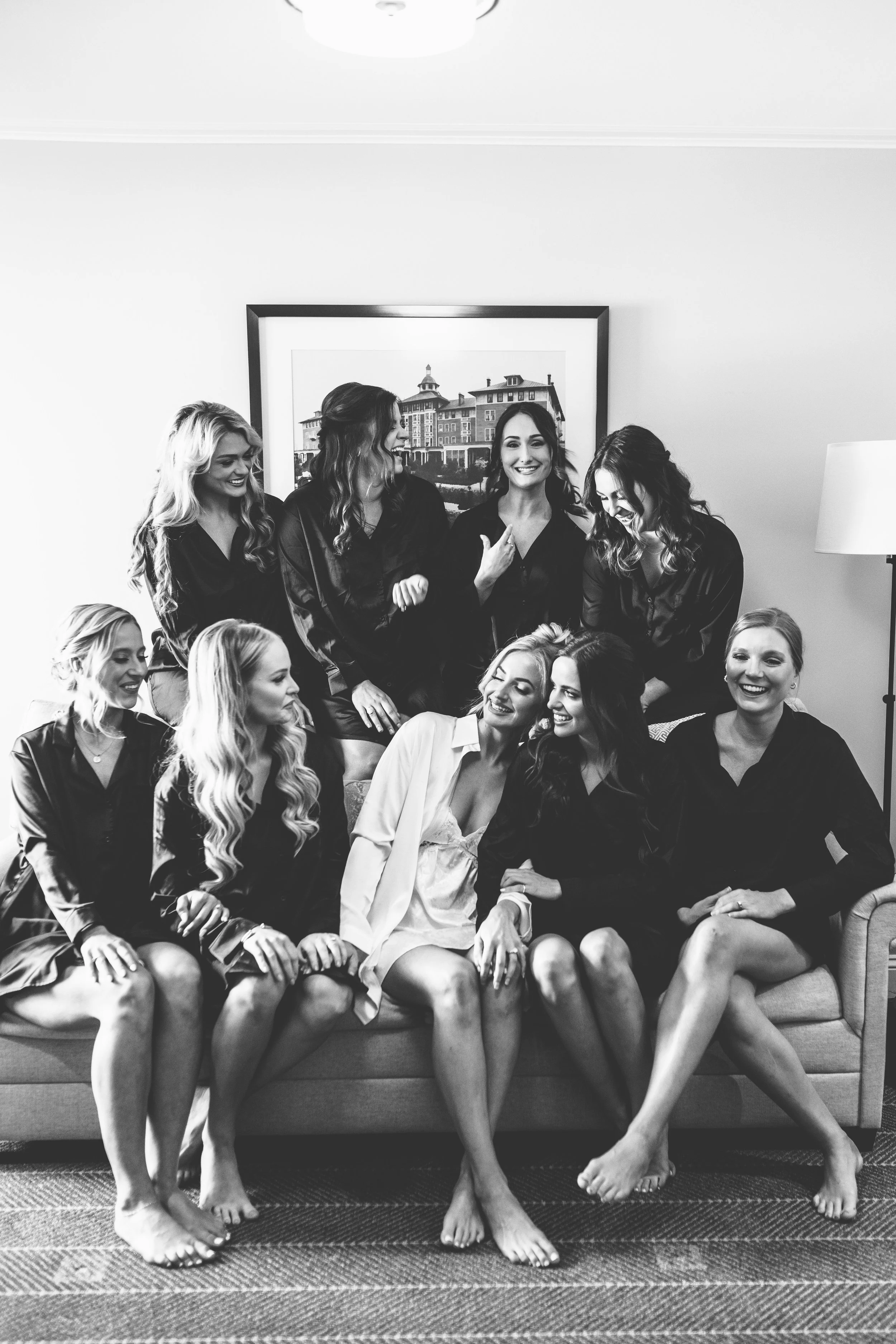 A group of nine women in pajamas sitting and standing on a sofa, smiling and laughing together in a living room with a framed picture on the wall behind them.