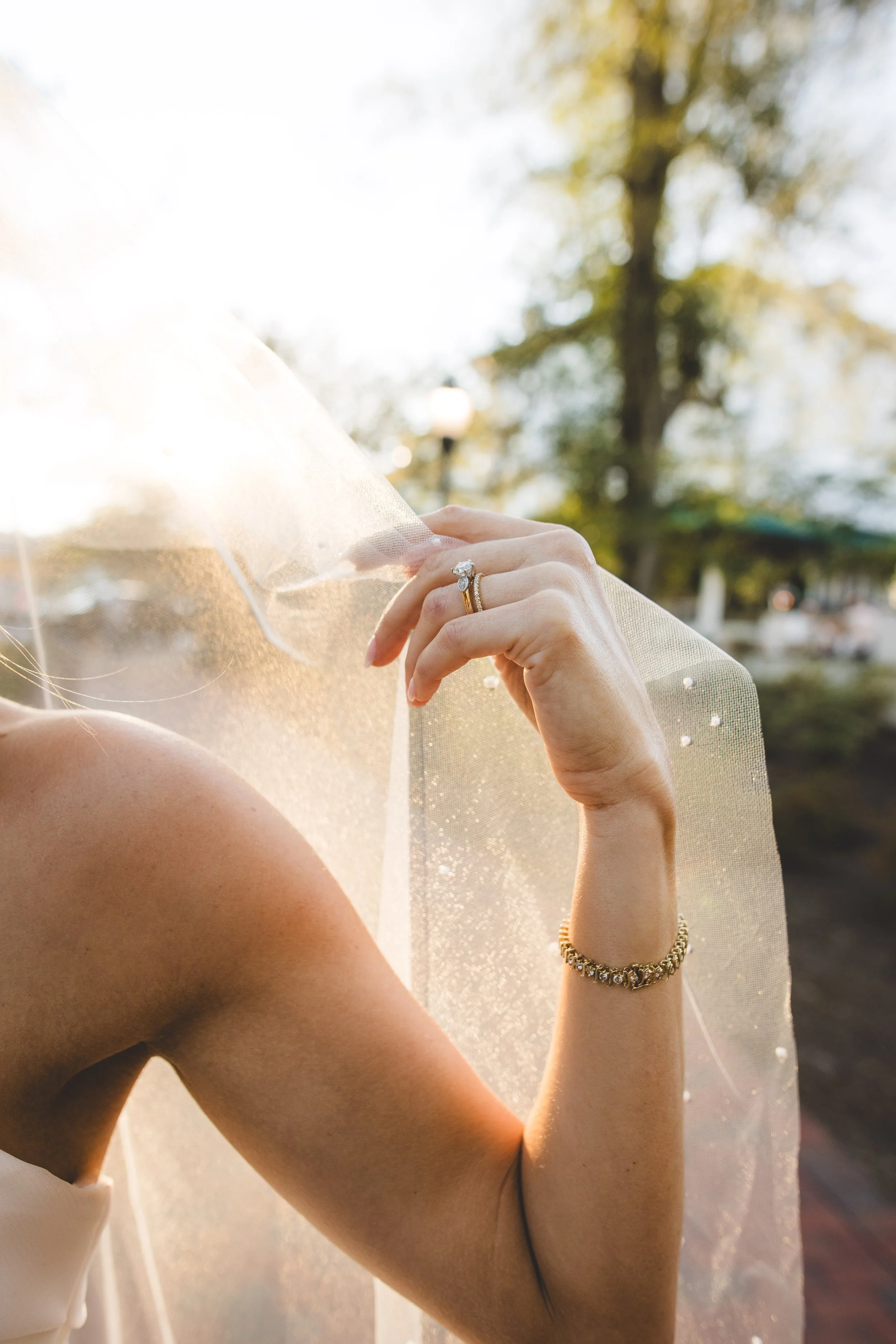 Close-up of a woman wearing an engagement ring on her ring finger, with sunlight backlighting her hand and veil. The background shows blurred trees and outdoor scenery.