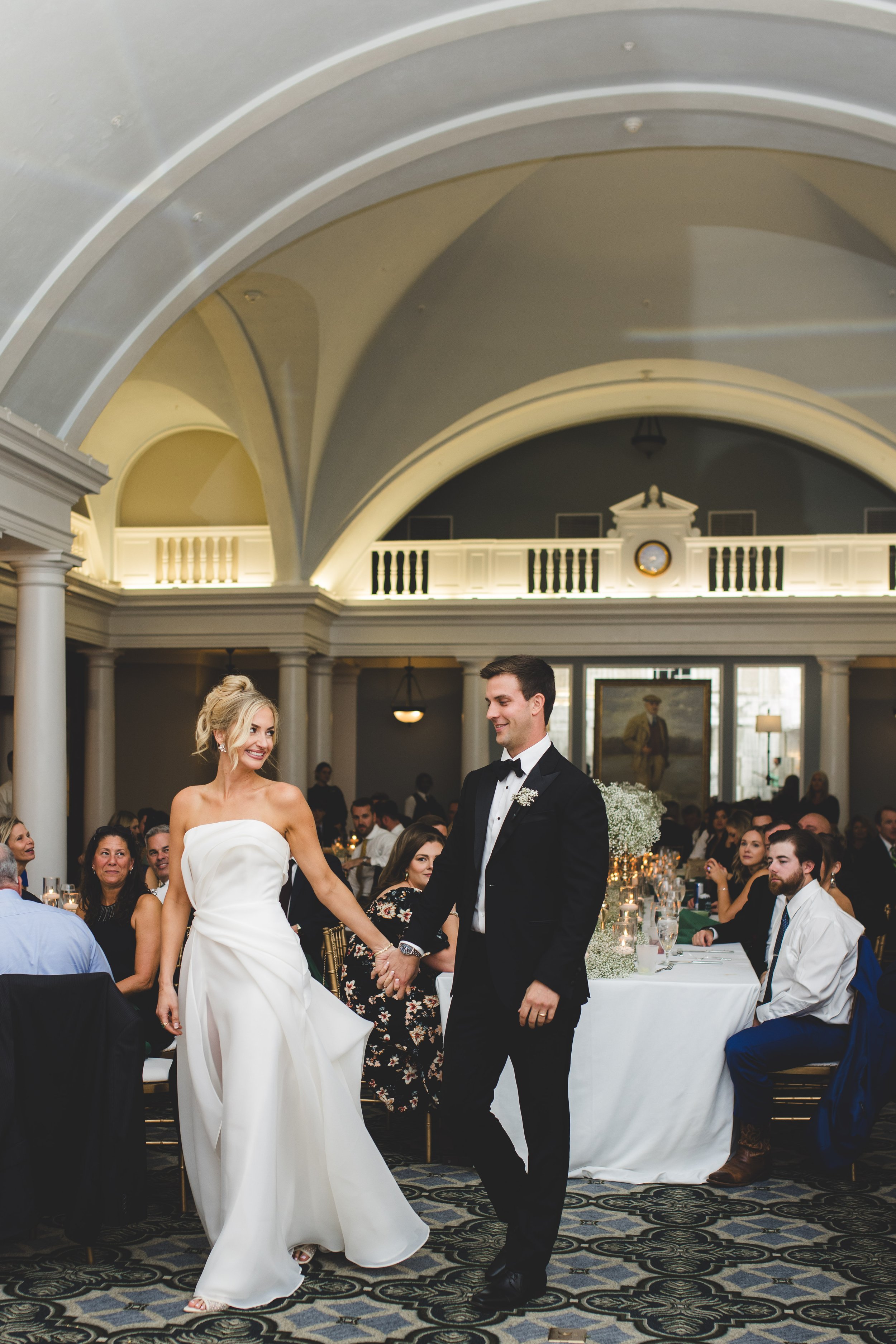 Bride and groom dancing at their wedding reception, surrounded by seated guests in an elegant hall with high arched ceilings.