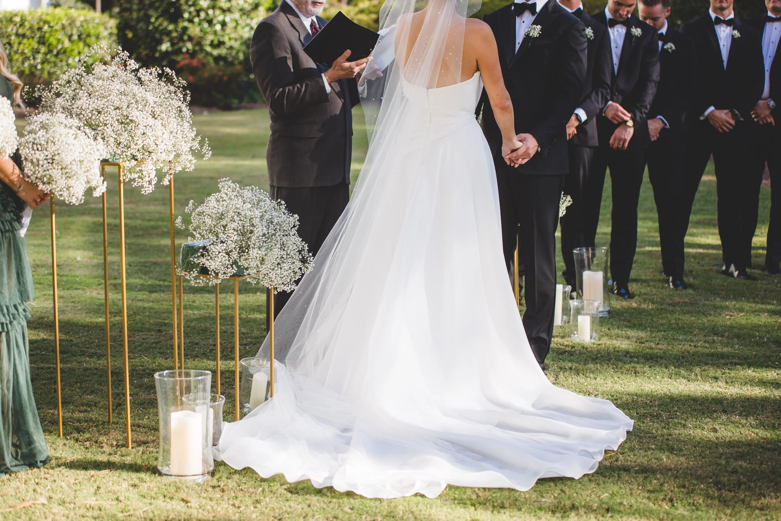 Bride and groom holding hands during an outdoor wedding ceremony with groomsmen, officiant, floral arrangements, and candles.