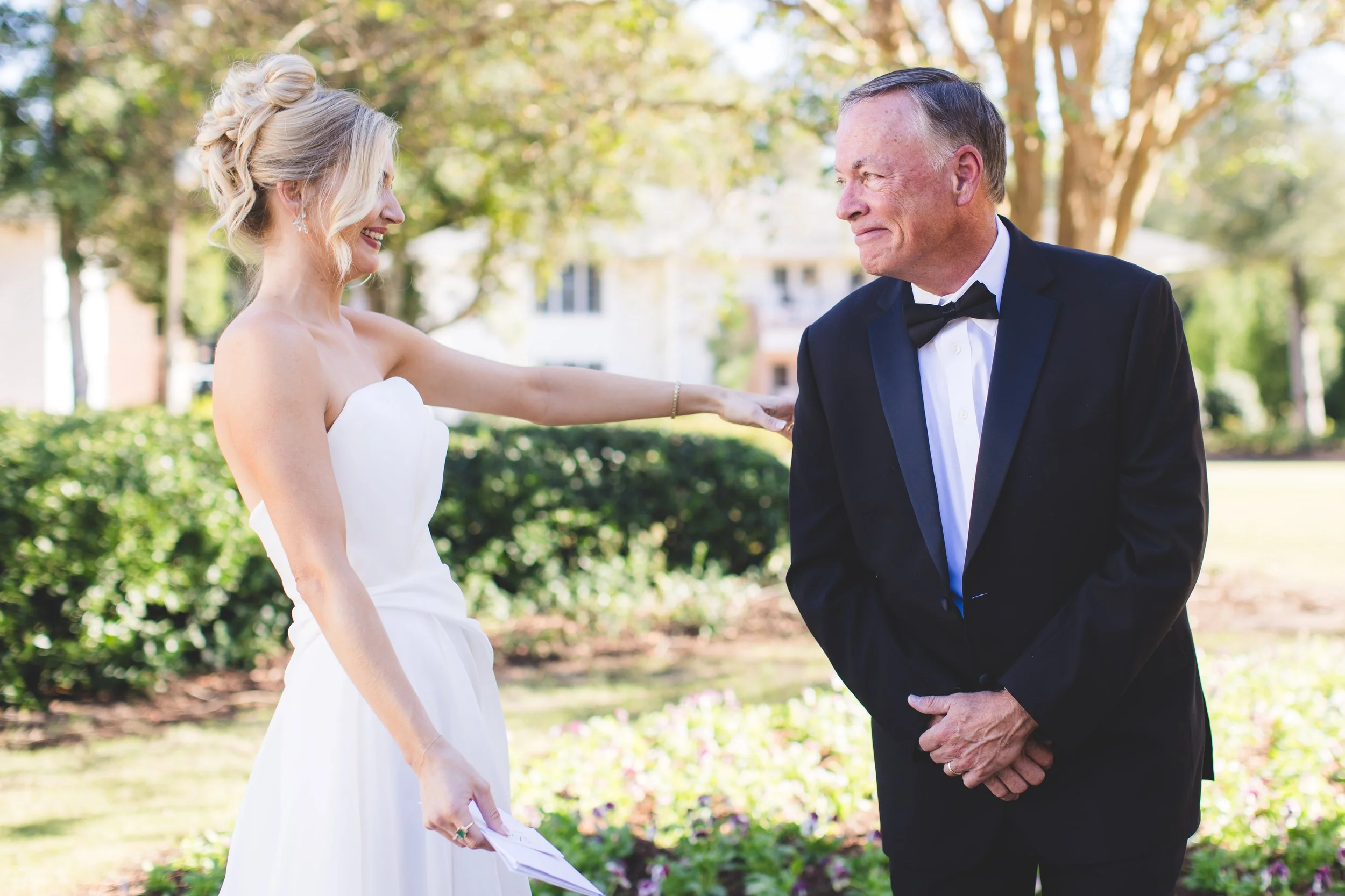 A bride in a white strapless wedding dress and a man in a tuxedo with a bow tie, outdoors with trees and houses in the background, smiling during a wedding ceremony.