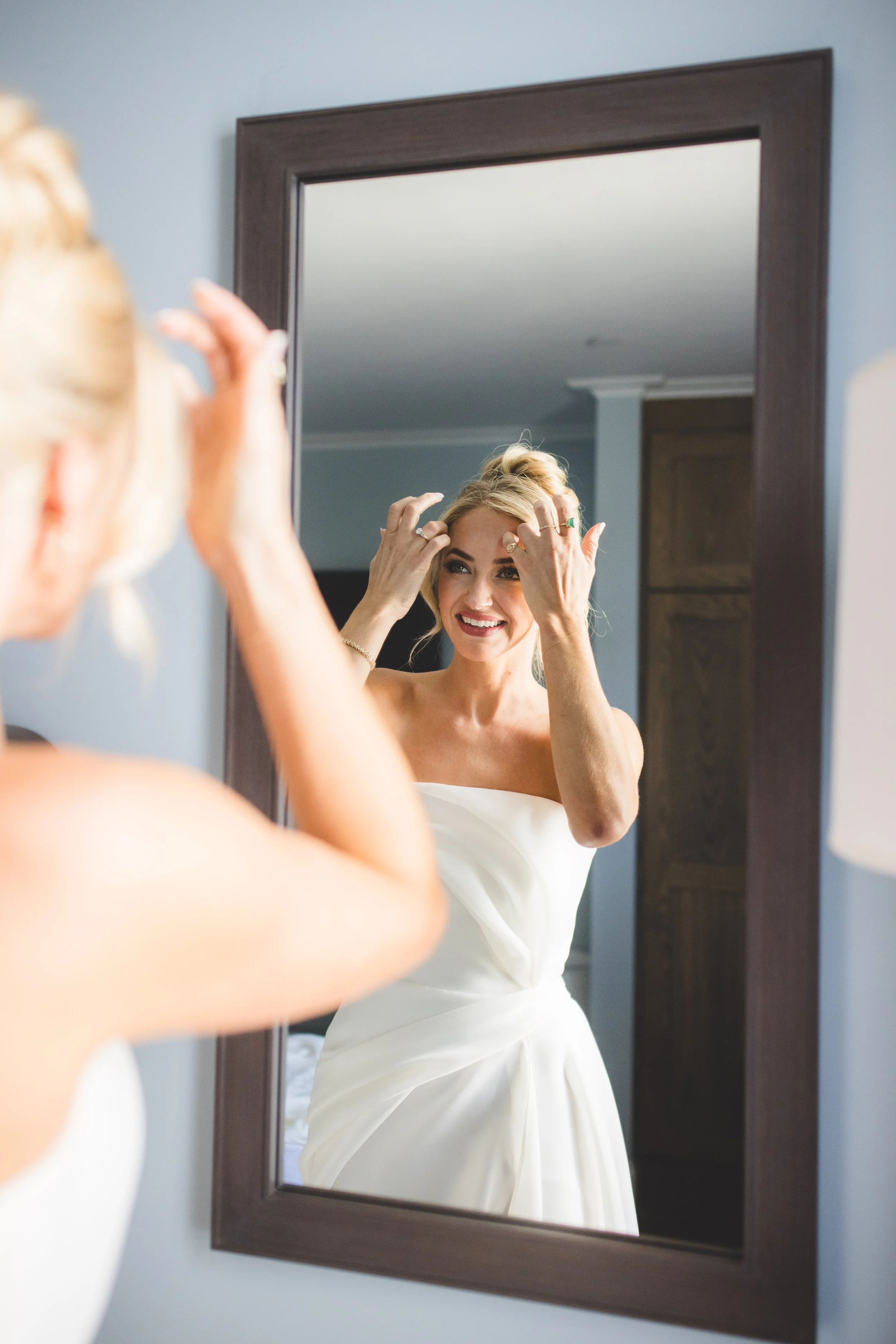 A smiling woman in a white dress looking at herself in the mirror, adjusting her hair.