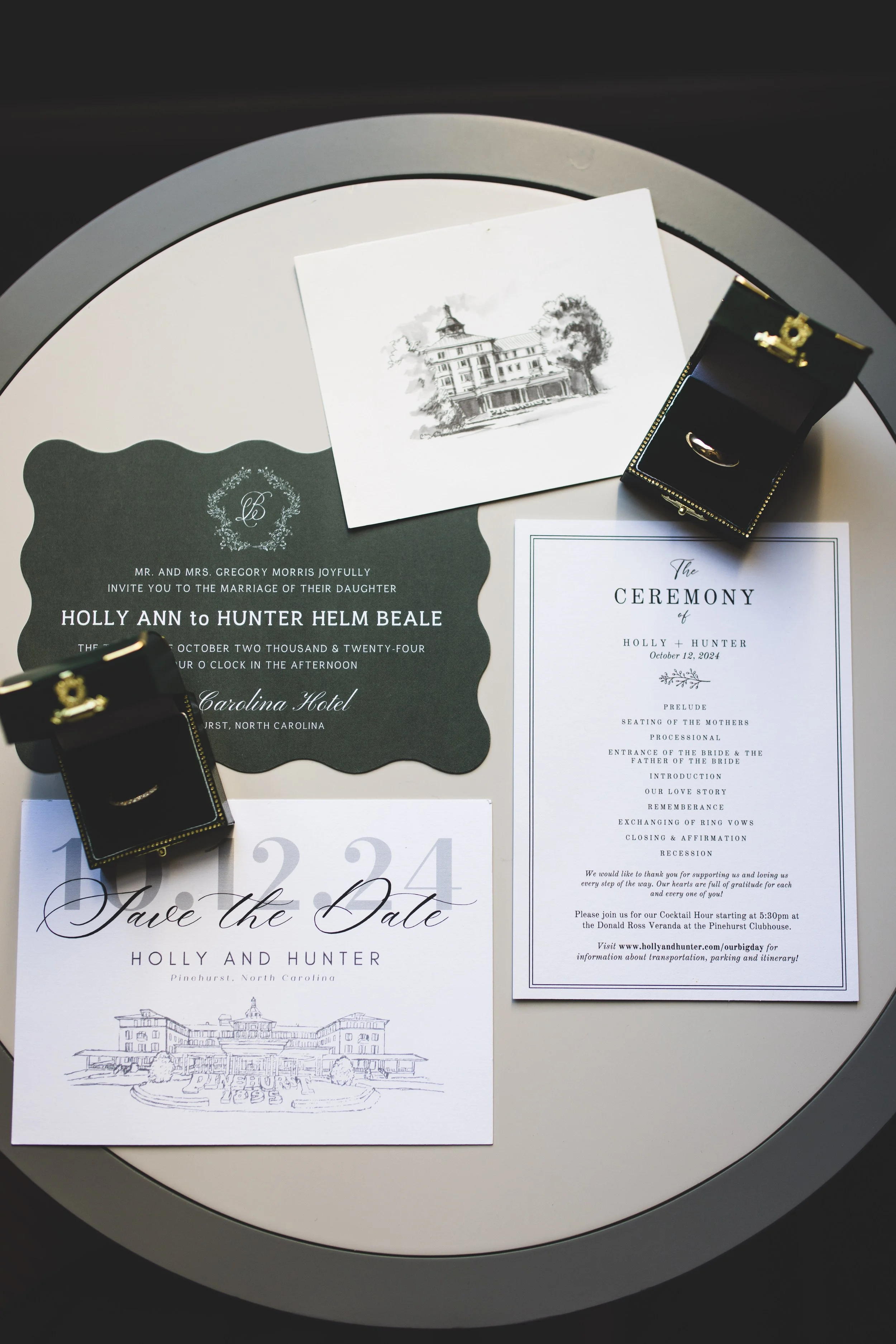 Wedding invitation suite on a round table, including an invitation card, a wedding program, a save the date card, and a small jewelry box with a ring, set against a black and white illustration of a building.