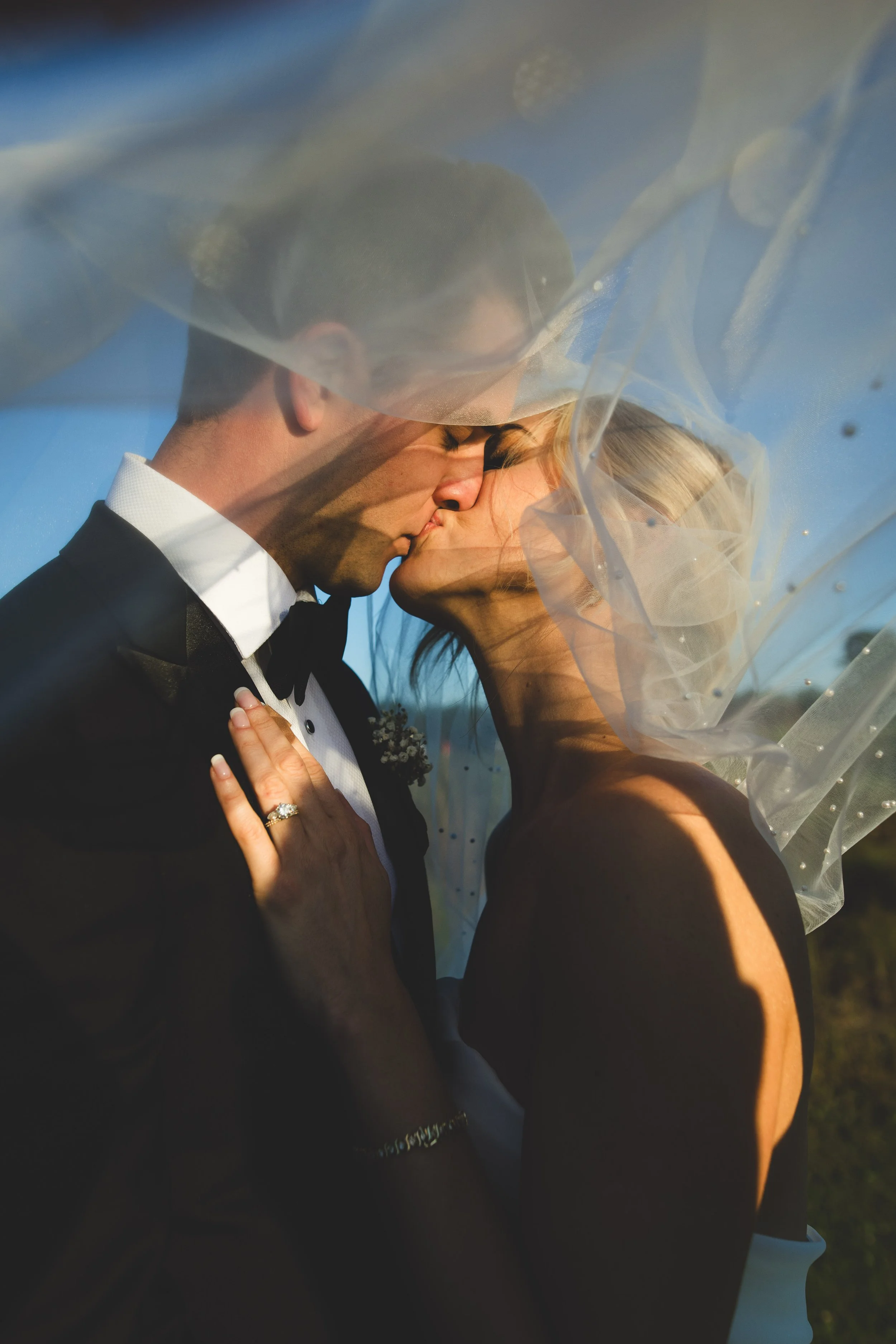 A bride and groom share a kiss under a veil, with soft sunlight illuminating their faces, set outdoors on a clear day.