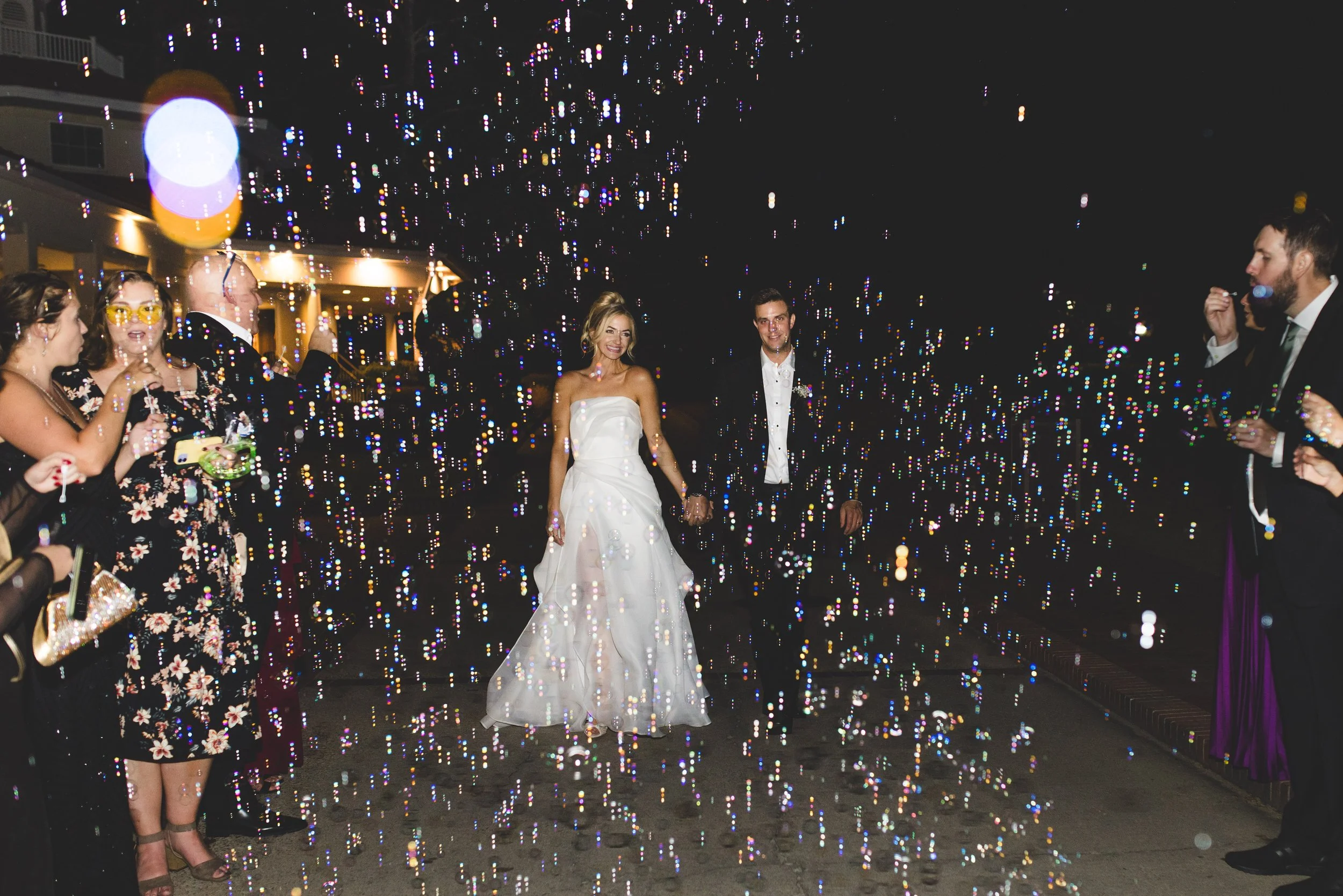 Bride and groom holding hands while walking through a shower of confetti at night, with guests on either side cheering and celebrating.