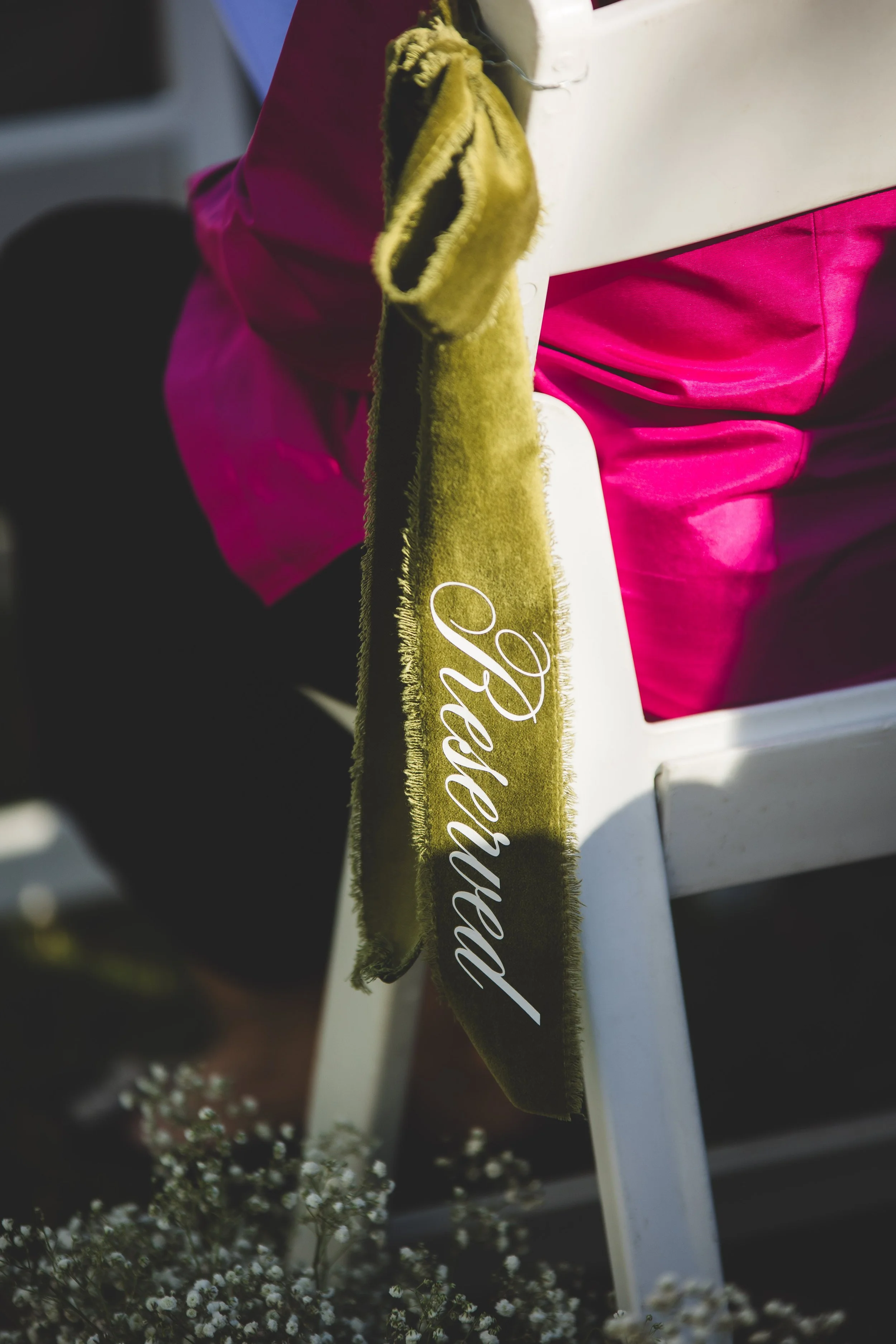 A white chair decorated with a green ribbon that reads "Bride" in white cursive letters, with pink fabric draped over the seat.