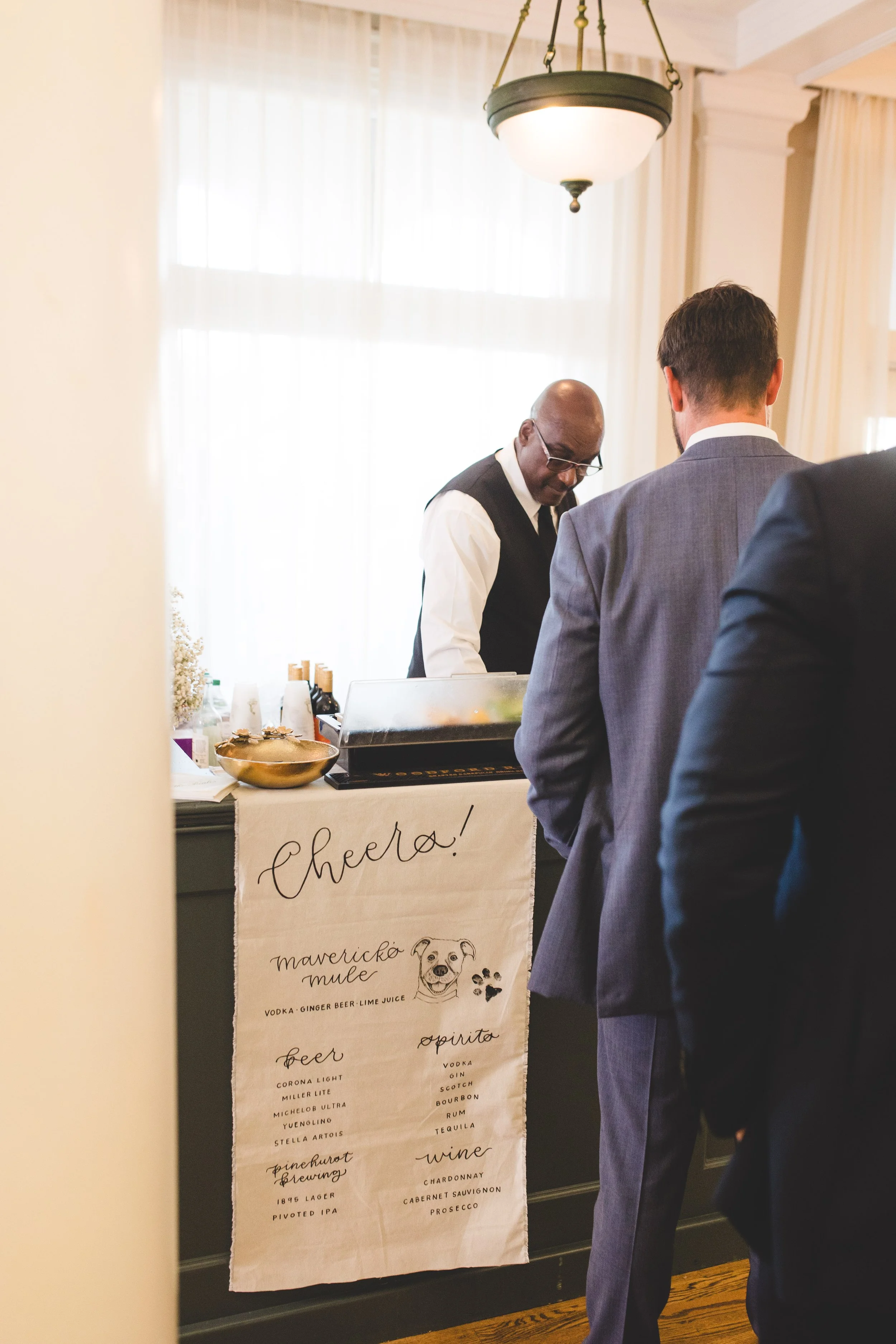 A bartender preparing drinks at a reception, with guests in formal attire. A sign on the bar says 'Cheers!' and lists beverages including beer, wine, and spirits.