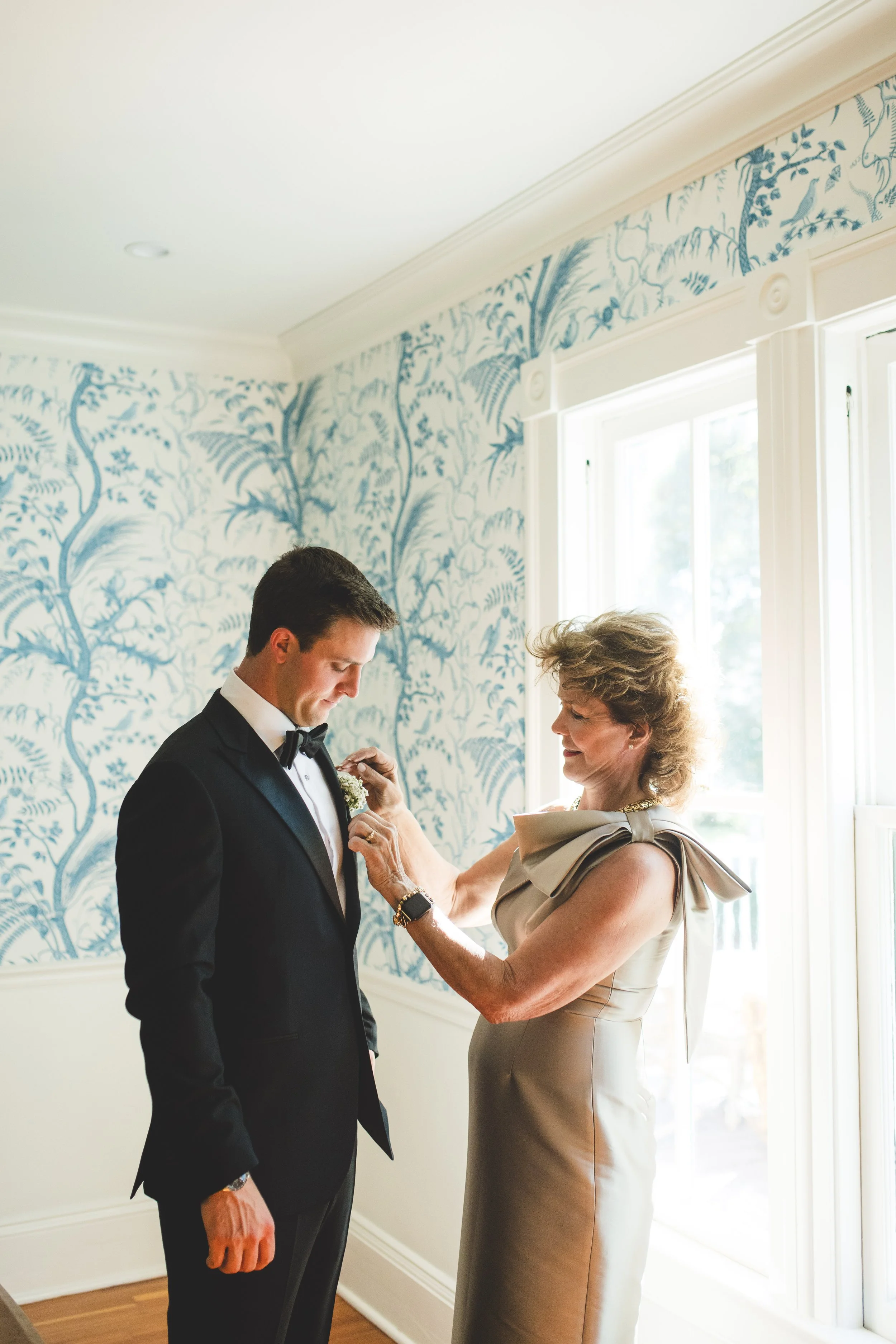 A woman in a beige dress pins a boutonniere on a man in a black tuxedo during a wedding preparation in a bright room with white and blue patterned wallpaper.