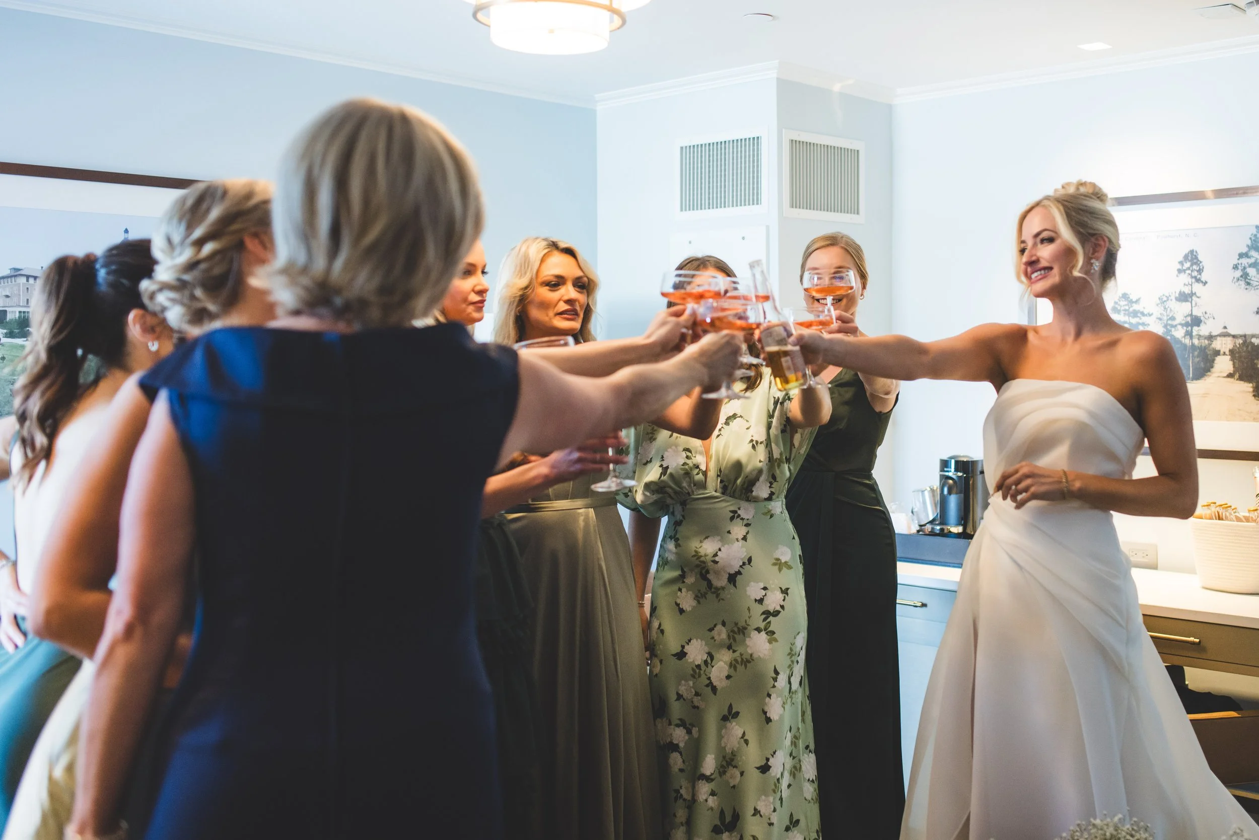 A group of women, including a bride in a white wedding dress, raising glasses in a toast during a celebration in a well-lit room.