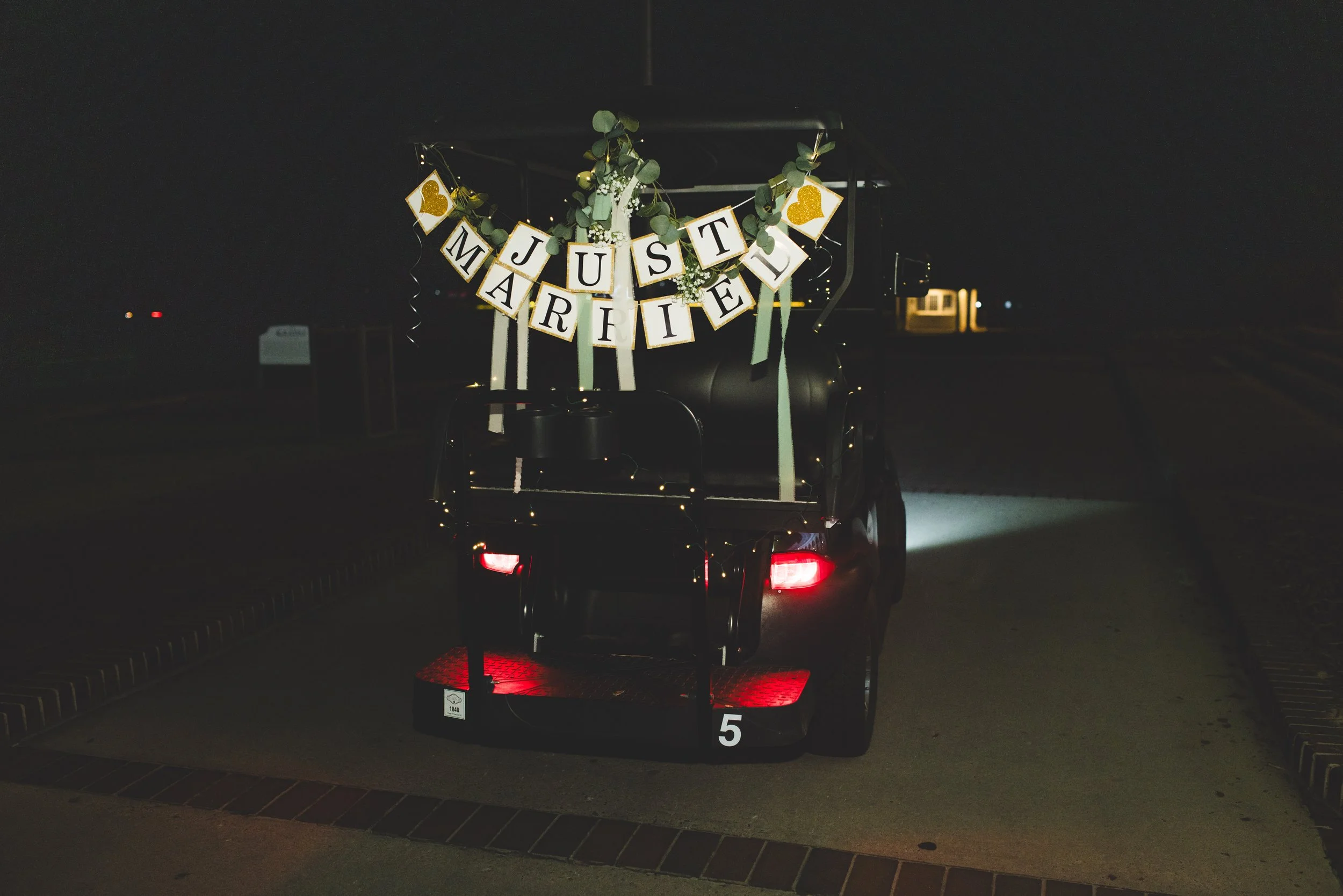 A golf cart decorated with a "Just Married" banner, greenery, and gold hearts, parked on a night street with minimal lighting.