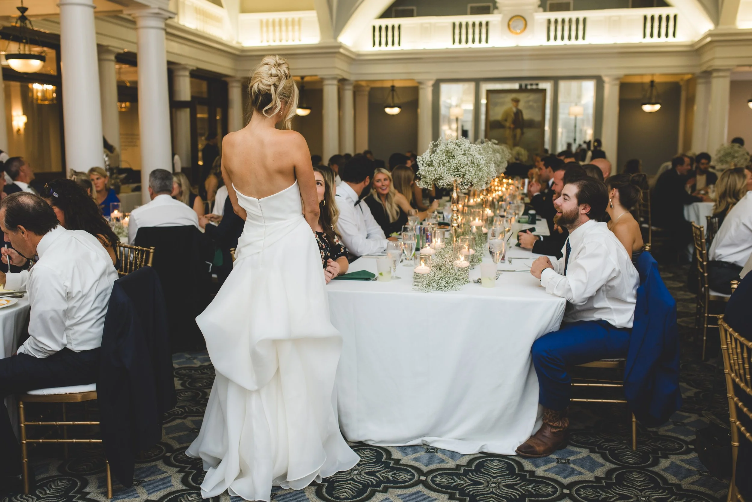 A woman in a strapless white wedding gown standing at a table, speaking to seated guests in a formal venue with white columns and elegant decor.