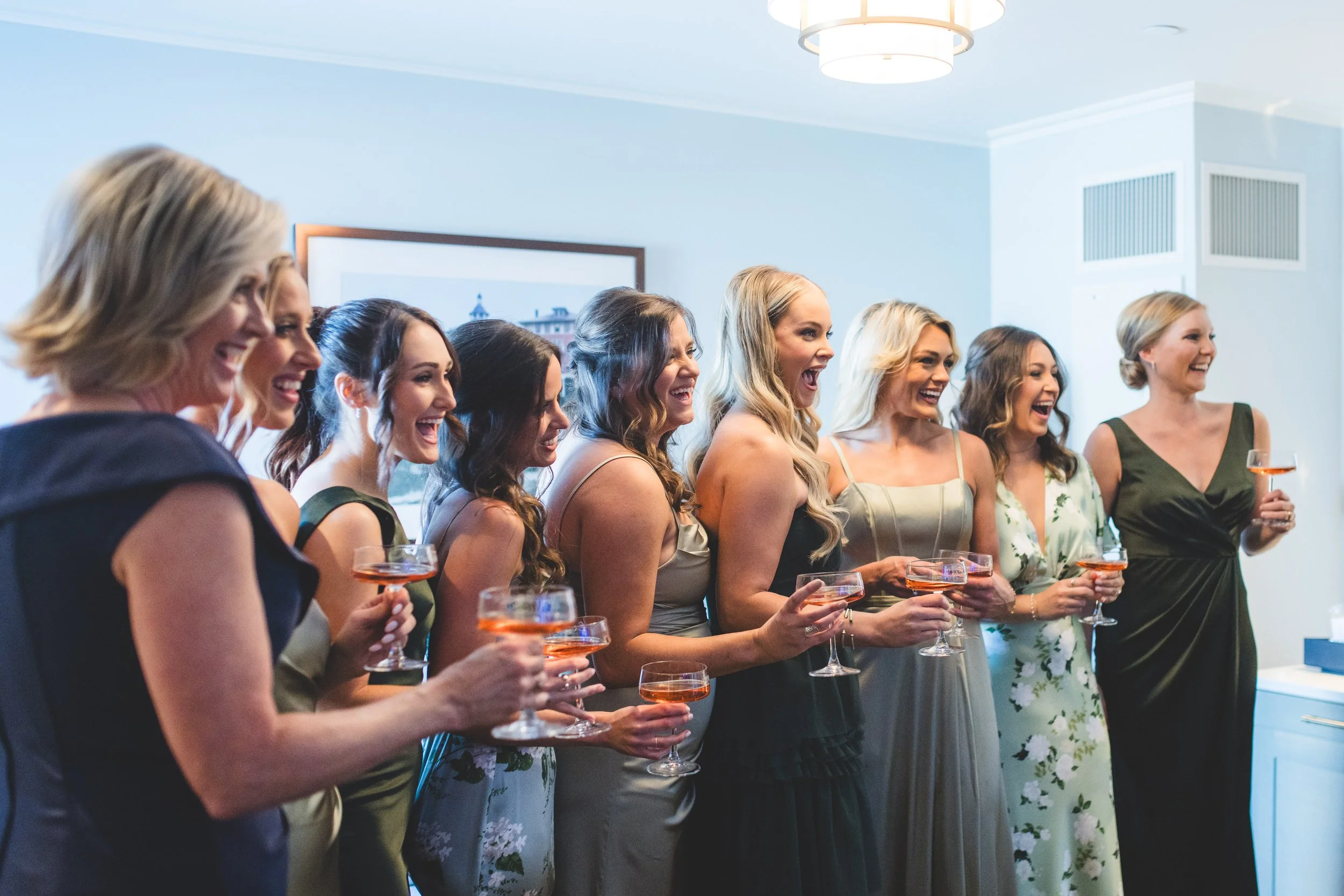 Group of nine women in formal dresses, holding glasses of rosé wine, smiling and laughing at a celebration or gathering indoors.