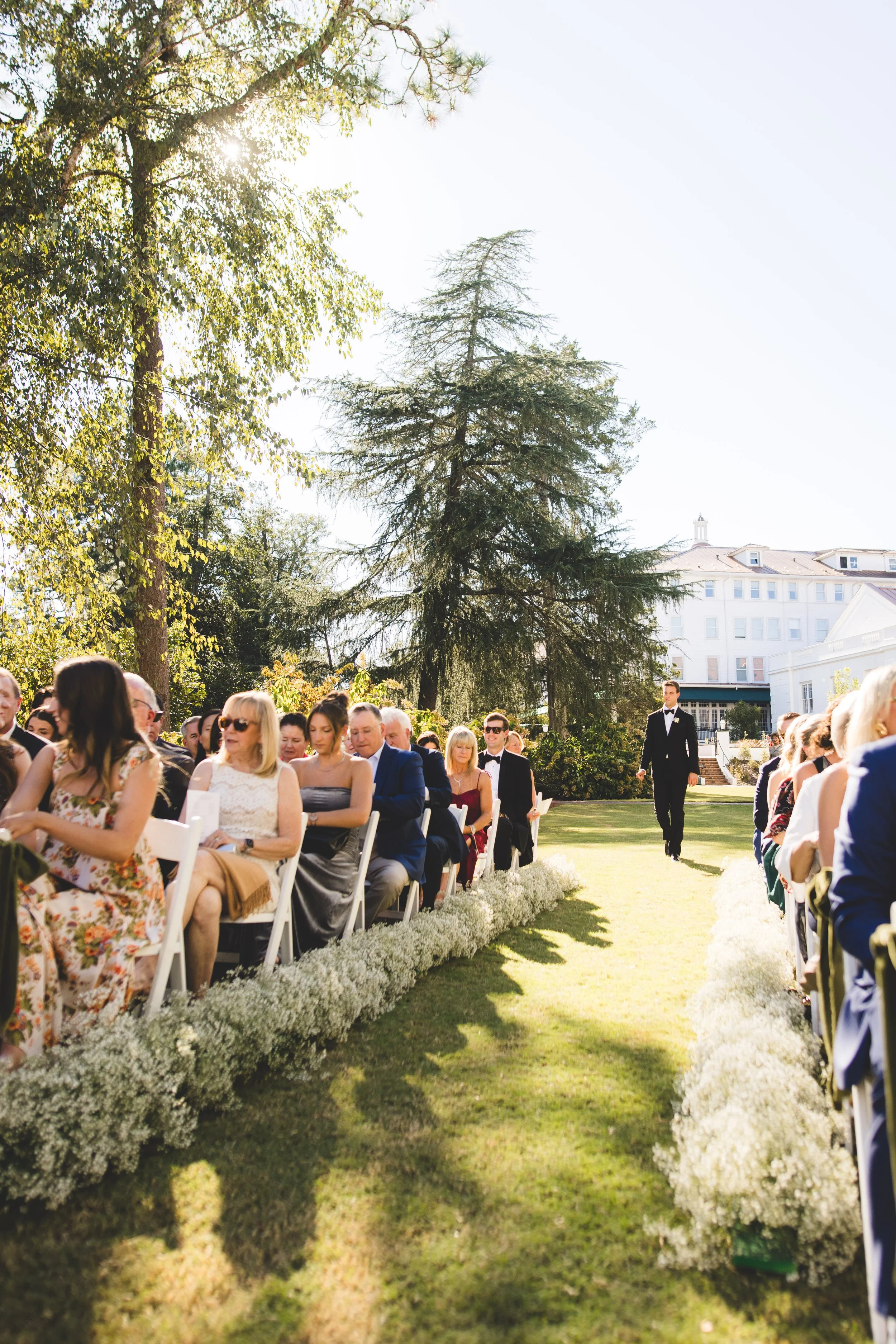 People attending an outdoor wedding ceremony on a sunny day, seated on white chairs along a grass aisle lined with white flowers, with trees and a large white building in the background.