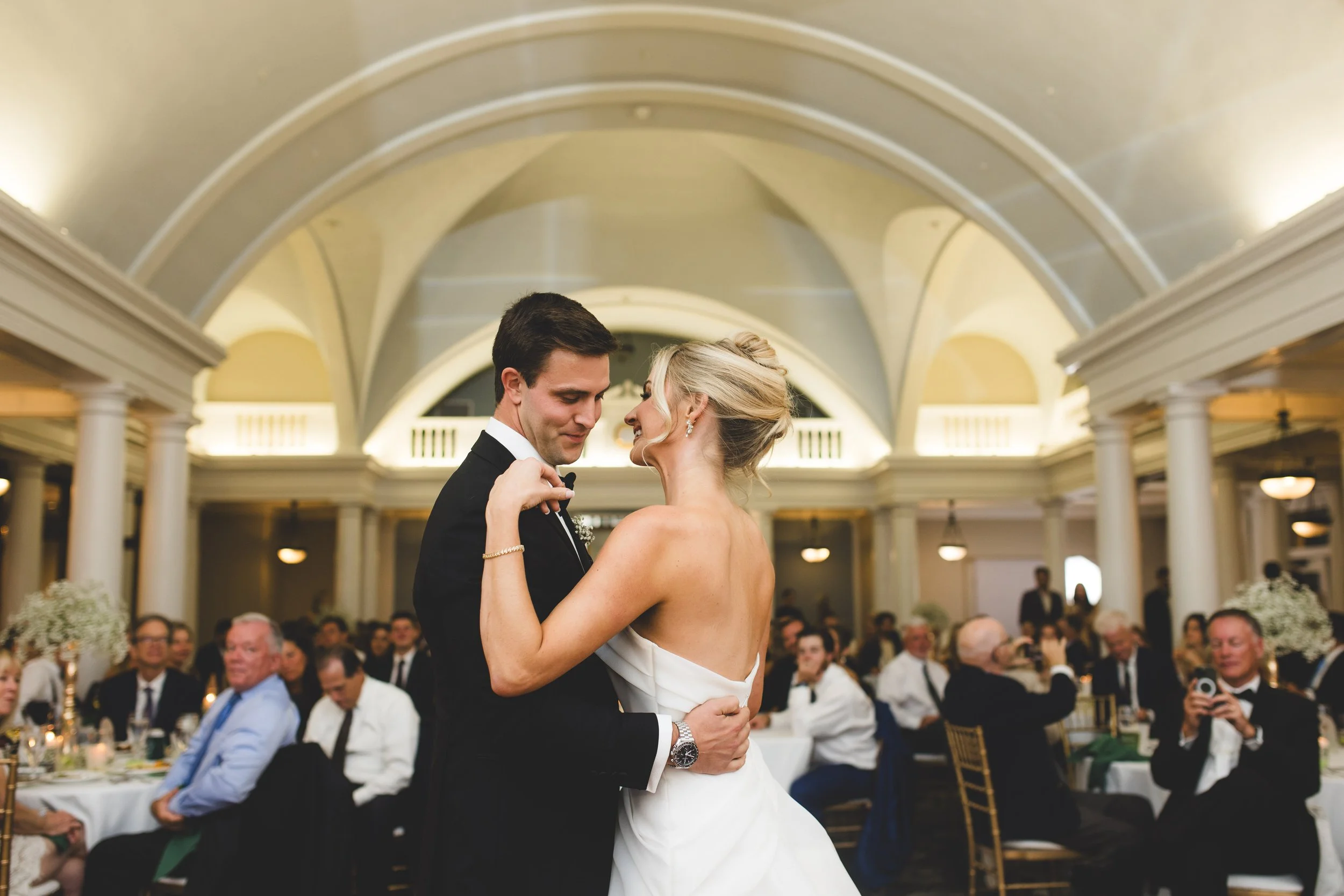 Bride and groom share a dance at their wedding reception in an elegant banquet hall with high arched ceilings and surrounded by seated guests.