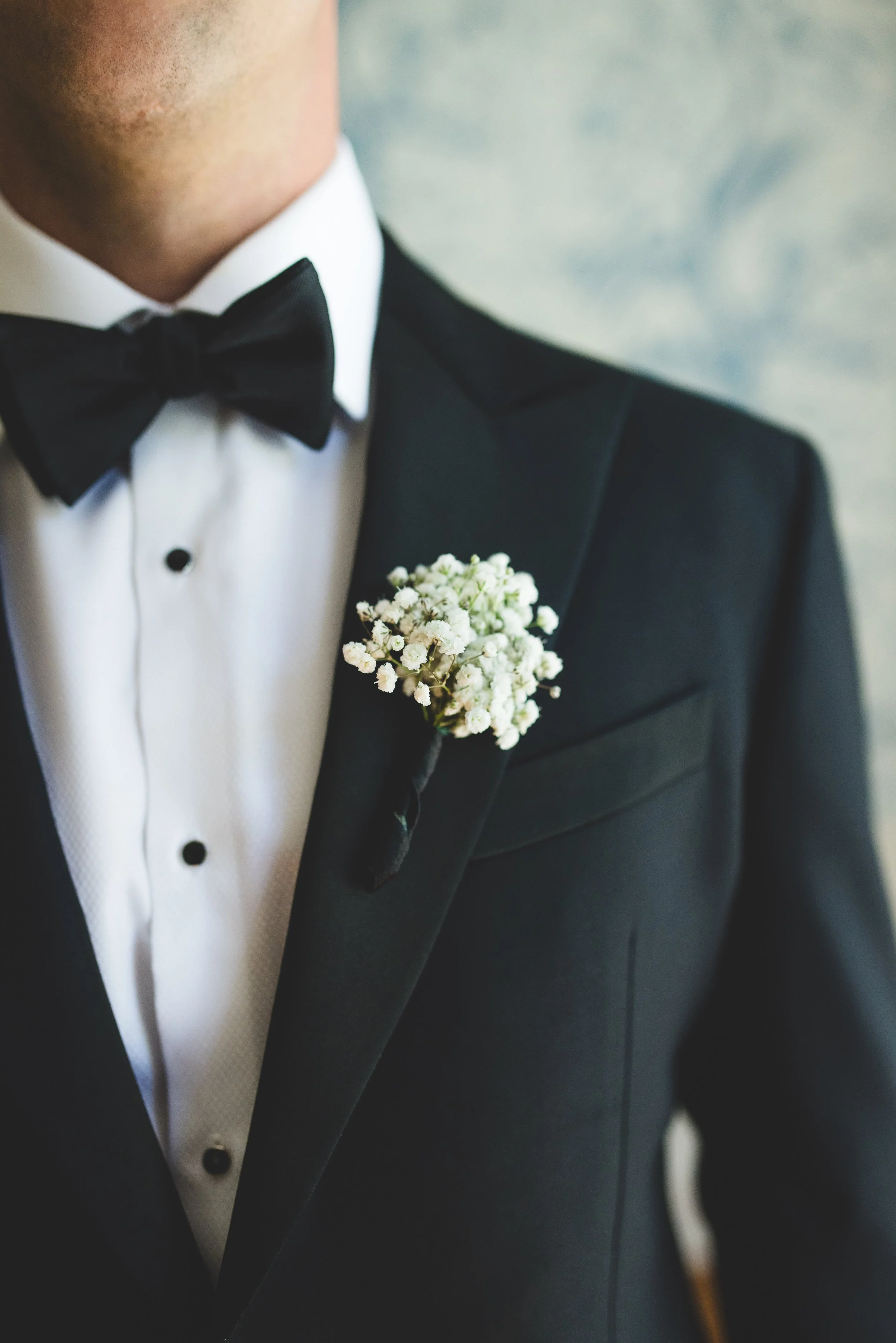 Close-up of a man in a tuxedo with a white dress shirt, black bow tie, and a white boutonniere on the lapel.