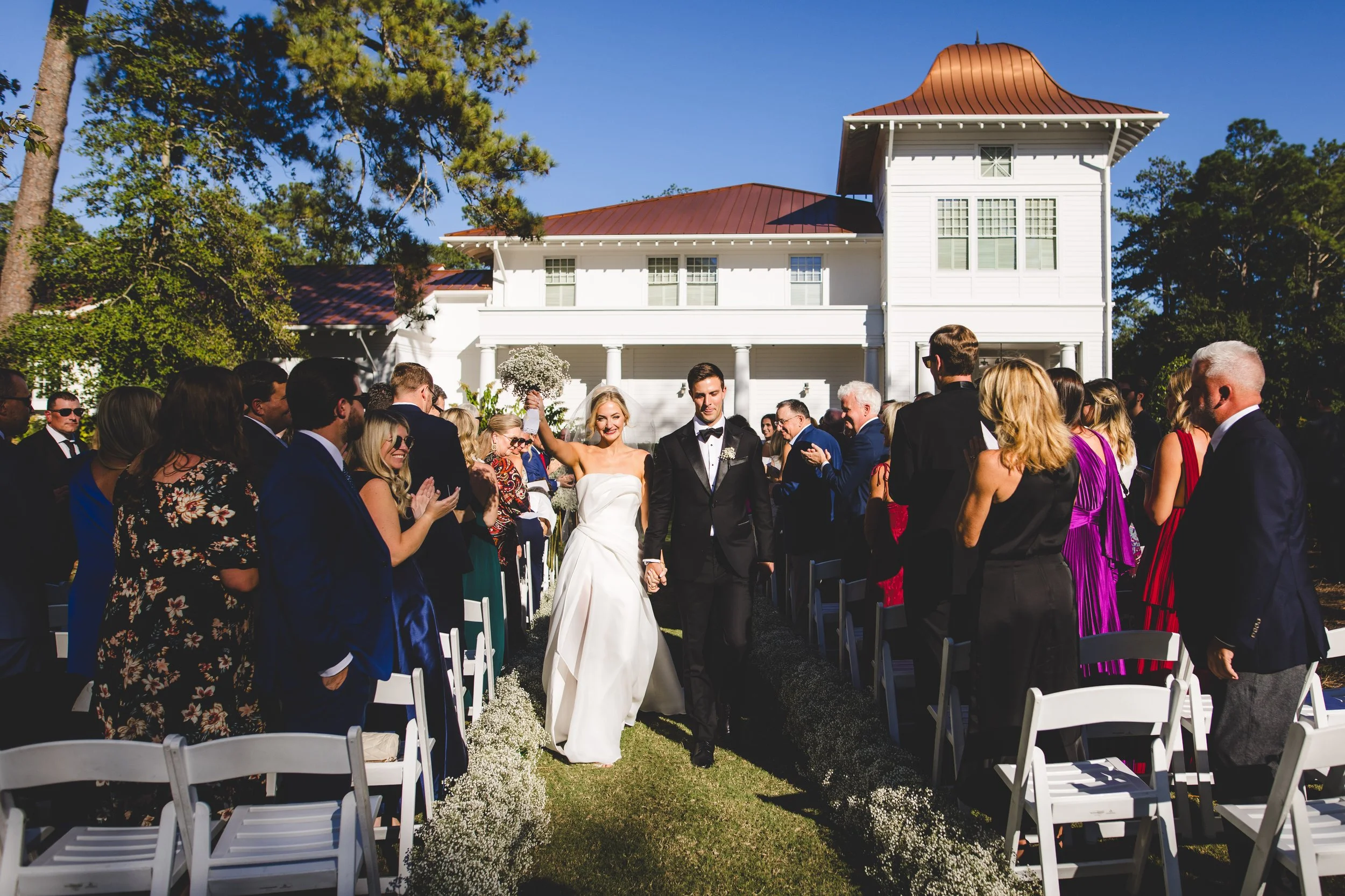 A newlywed couple walks down the aisle at an outdoor wedding ceremony, holding hands and smiling. Guests on either side of the aisle are clapping and cheering. The scene is set in front of a large white house with a red roof, under a clear blue sky, 