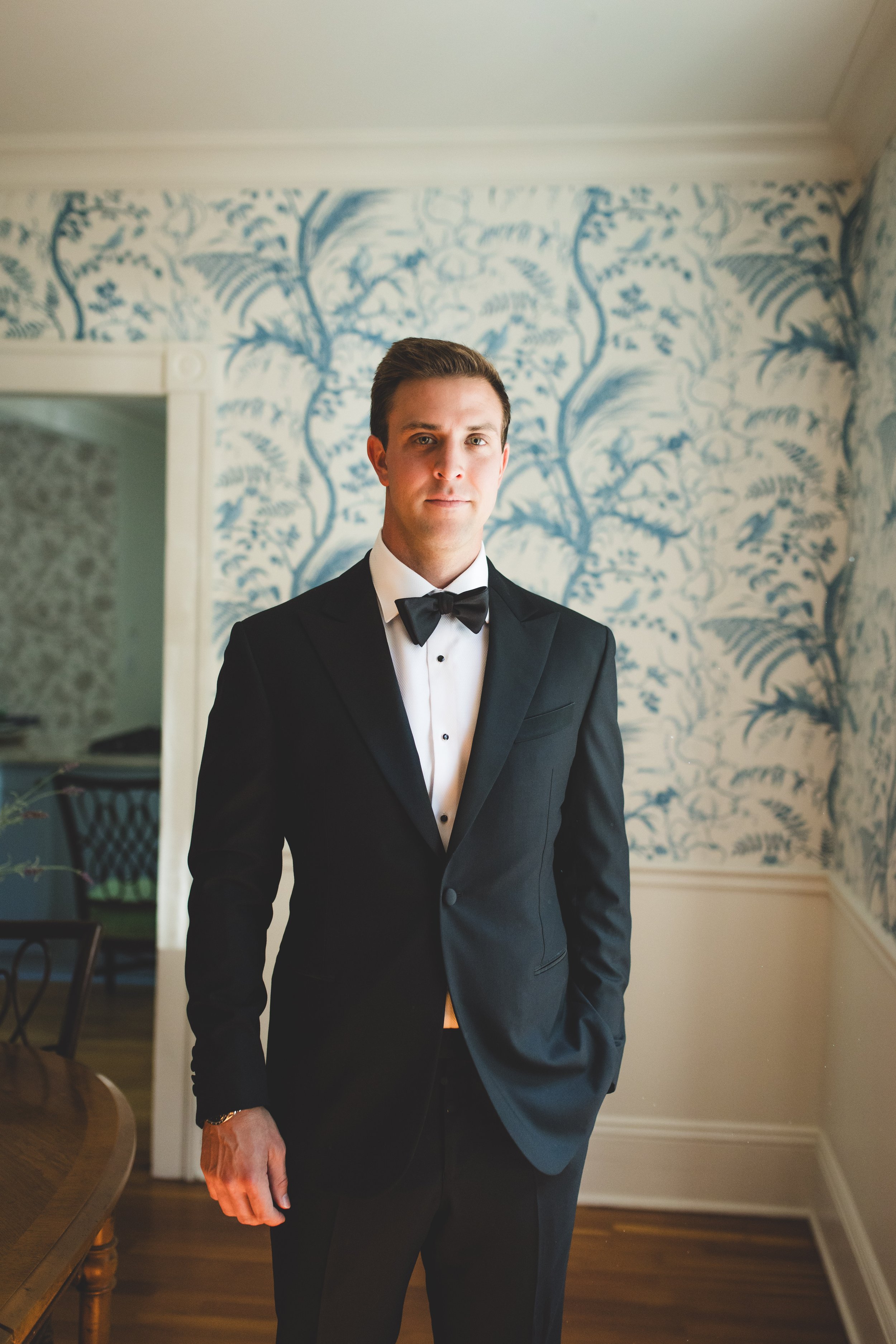 A well-dressed man in a black tuxedo with a bow tie standing indoors, with a blue and white patterned wallpaper in the background.