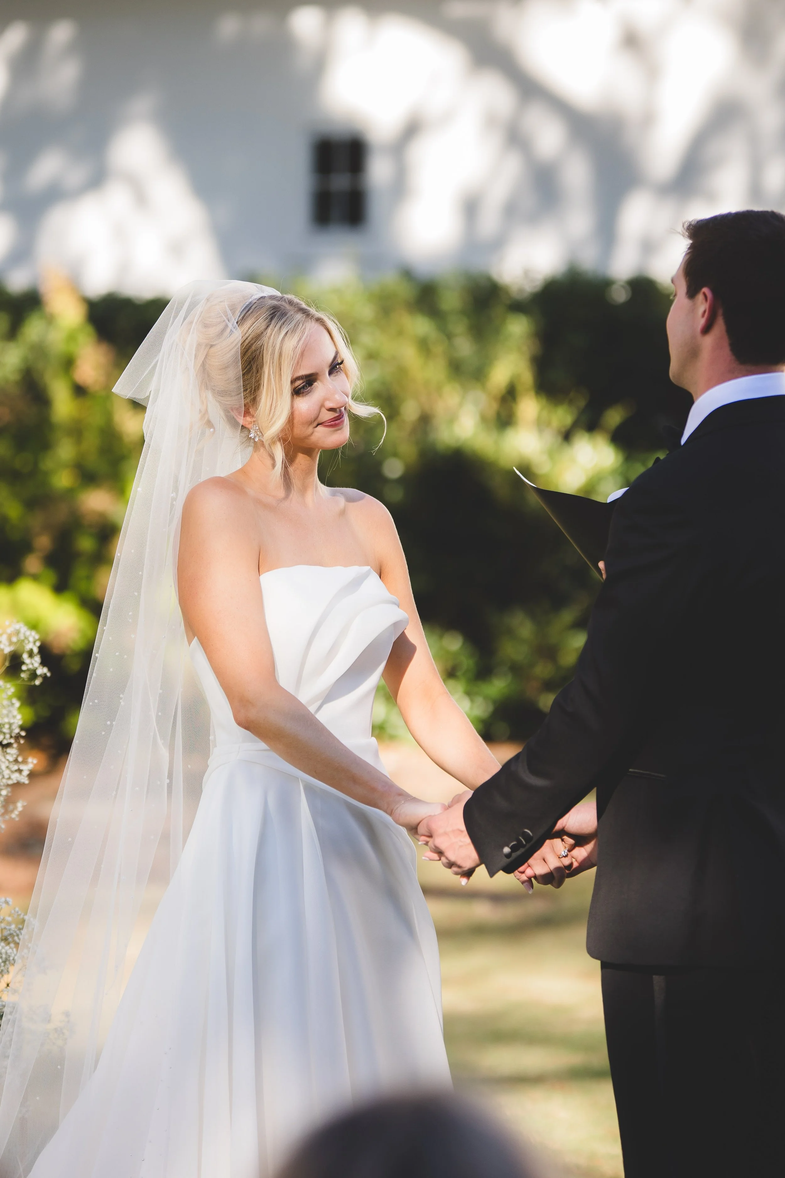 A bride and groom holding hands during their outdoor wedding ceremony, facing each other, with a white house and greenery in the background.