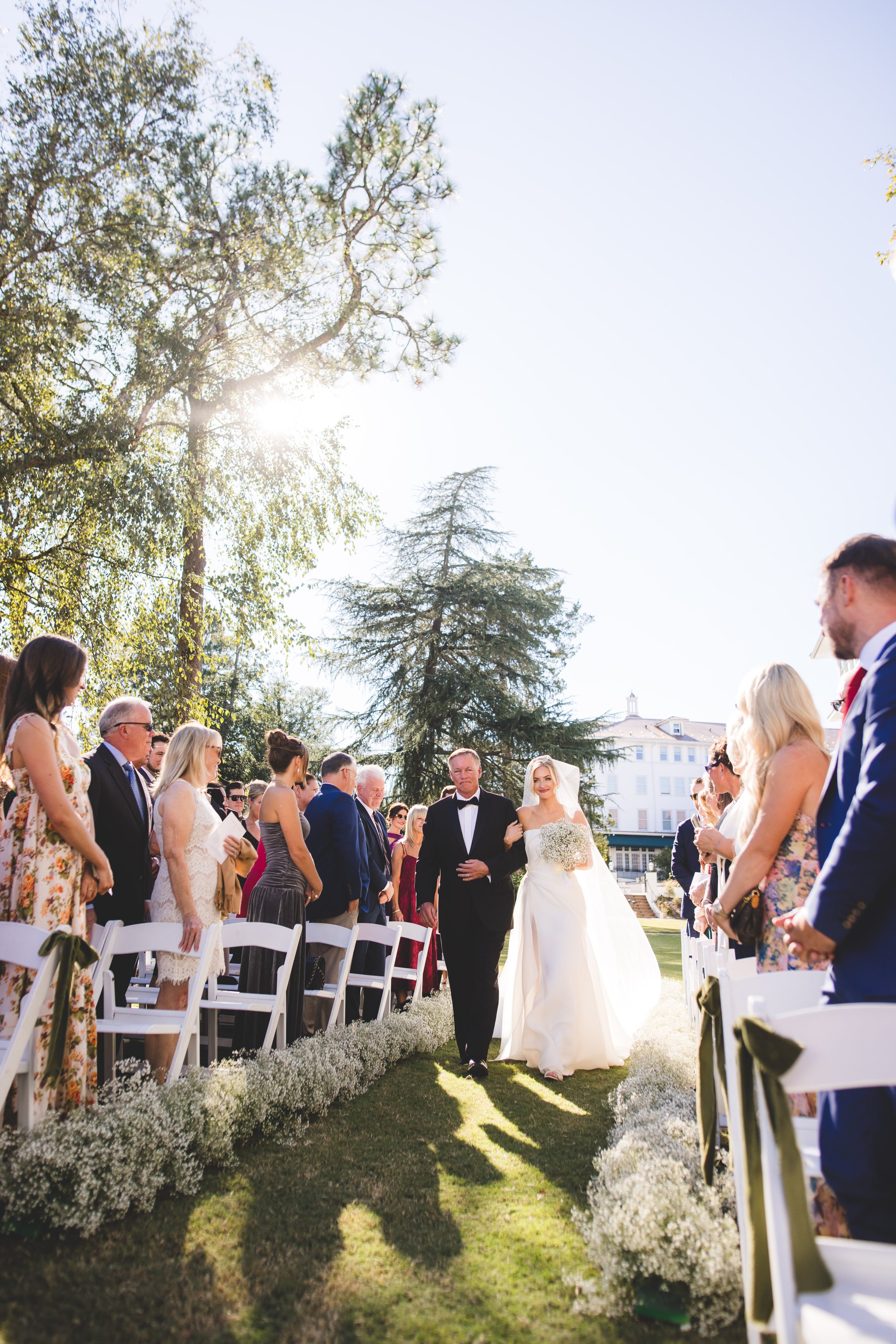 Bride walking down the aisle with her father at an outdoor wedding ceremony on a sunny day, with guests seated on both sides.
