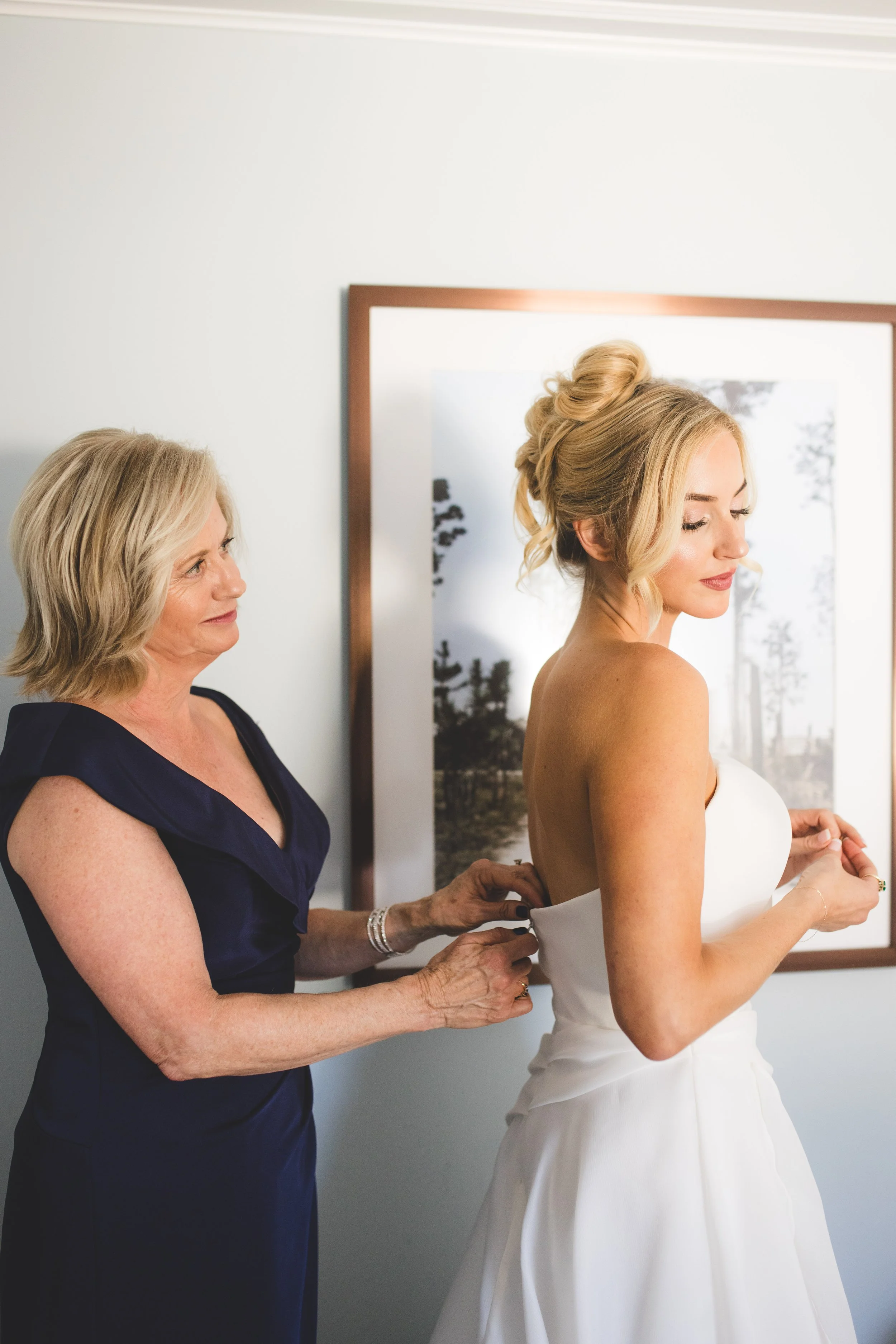A woman in a white wedding dress has her back to a woman in a dark dress, who is helping her adjust the dress.