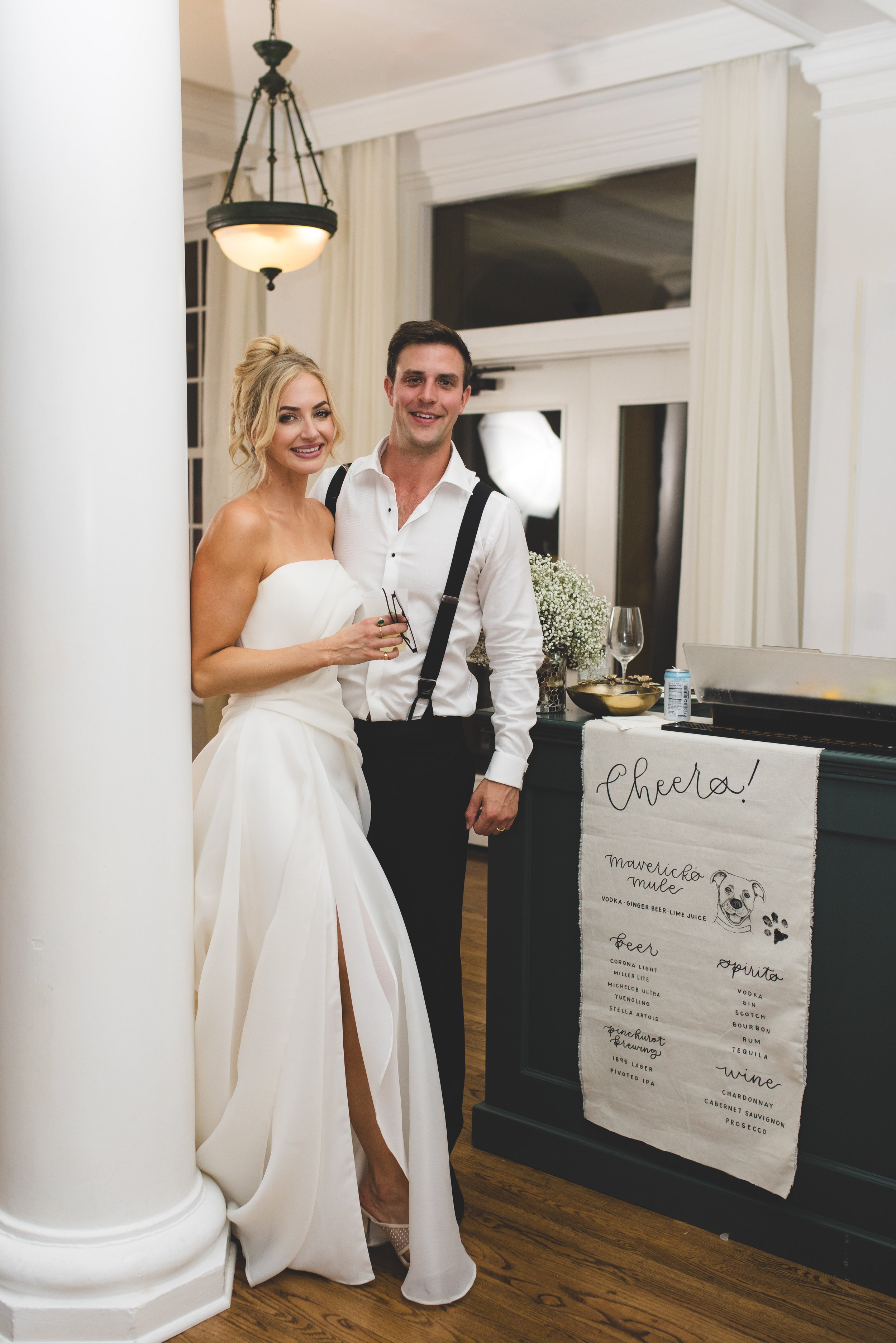 A bride in a white wedding gown and a groom in a white shirt and black suspenders standing together at a wedding reception, smiling, with a bar and a menu board nearby.