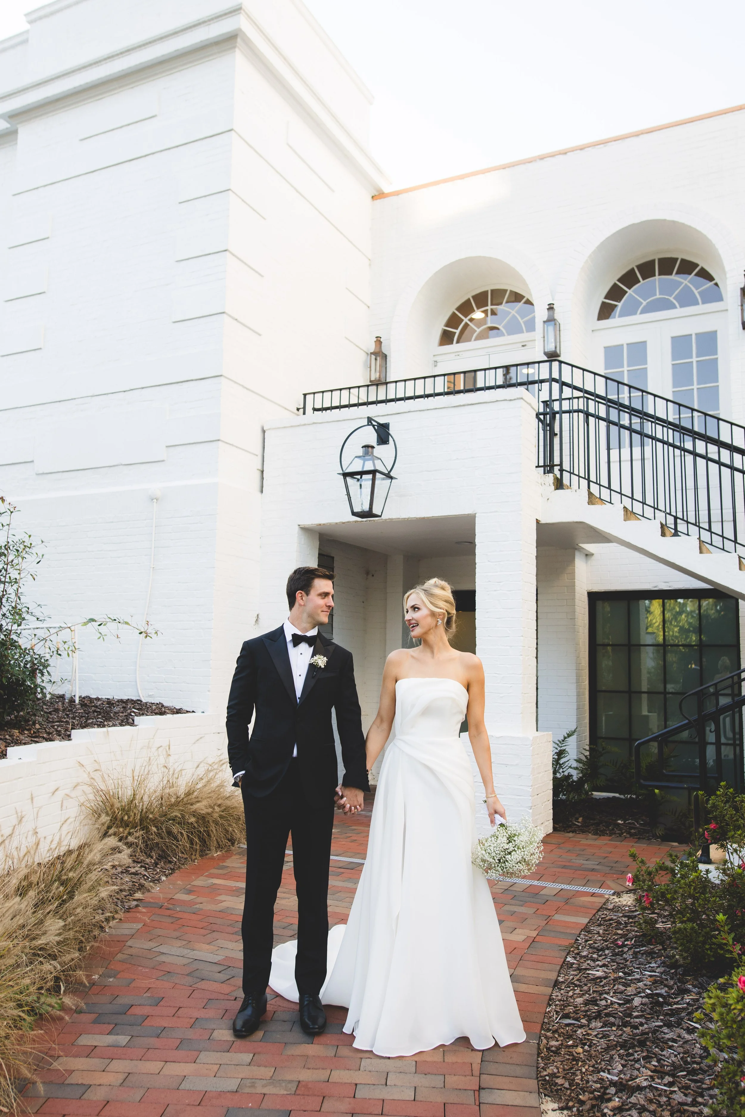 Bride and groom holding hands outside a white building, smiling at each other, bride in a strapless wedding dress holding a bouquet, groom in a black tuxedo, brick pathway with landscaping.