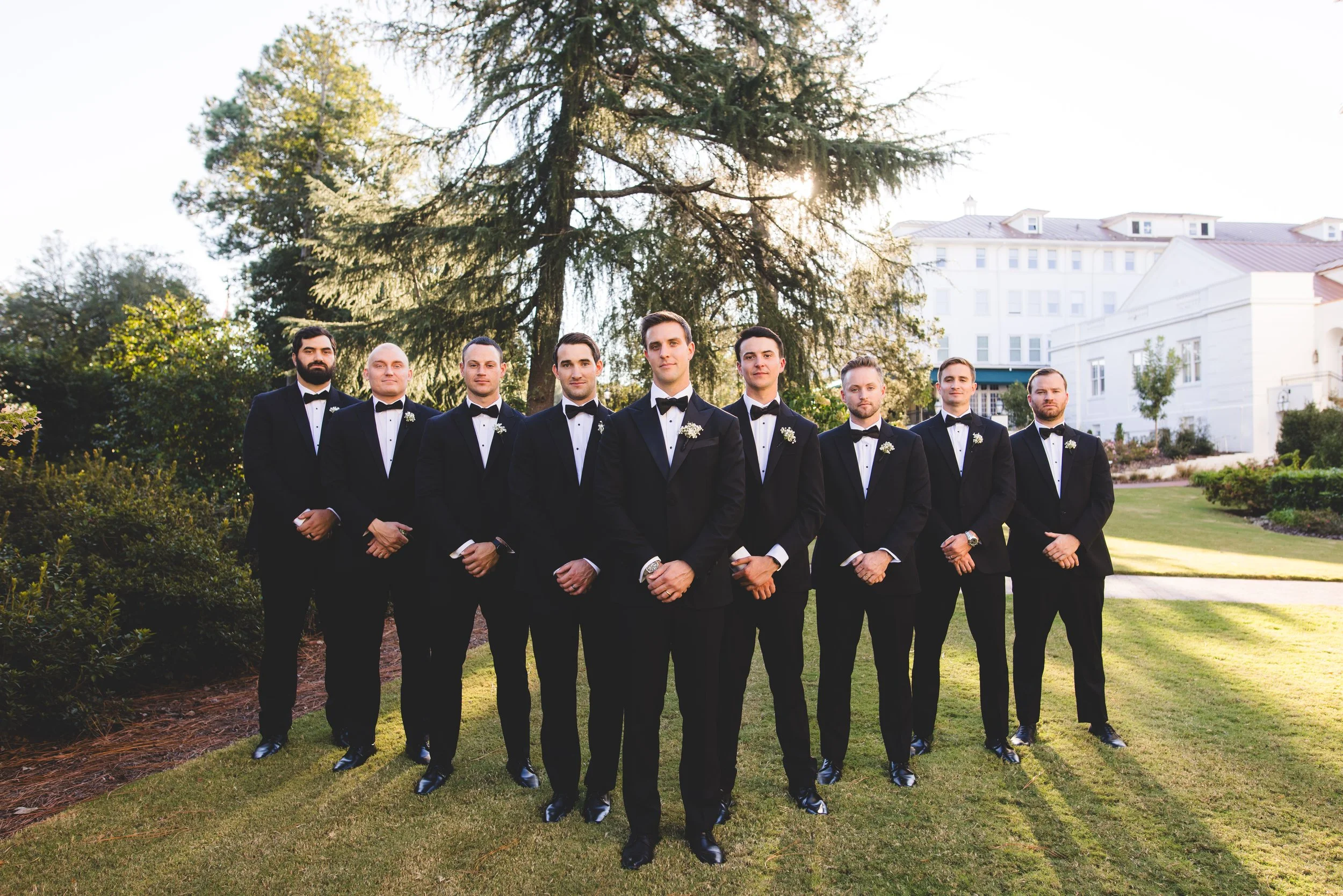 A group of eight men in black tuxedos with bow ties, standing outdoors on a sunny day, with large trees and a white building in the background.