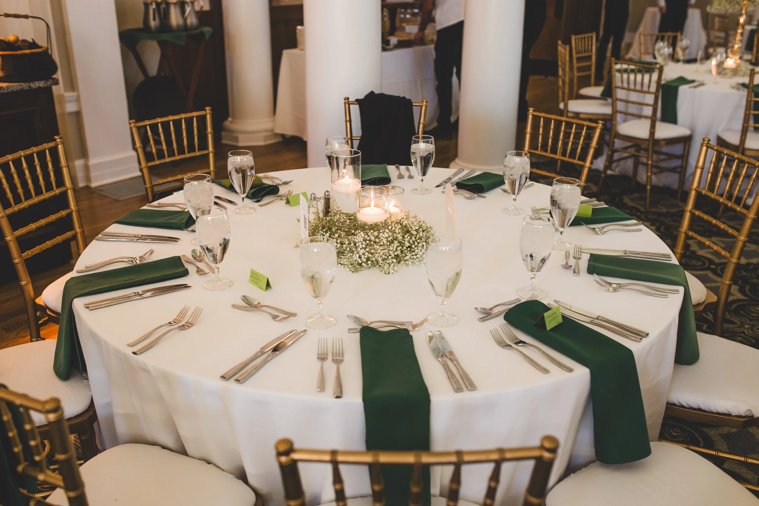 Round banquet table with white tablecloth, gold chairs, green napkins, glassware, and candle centerpiece in a decorated event hall