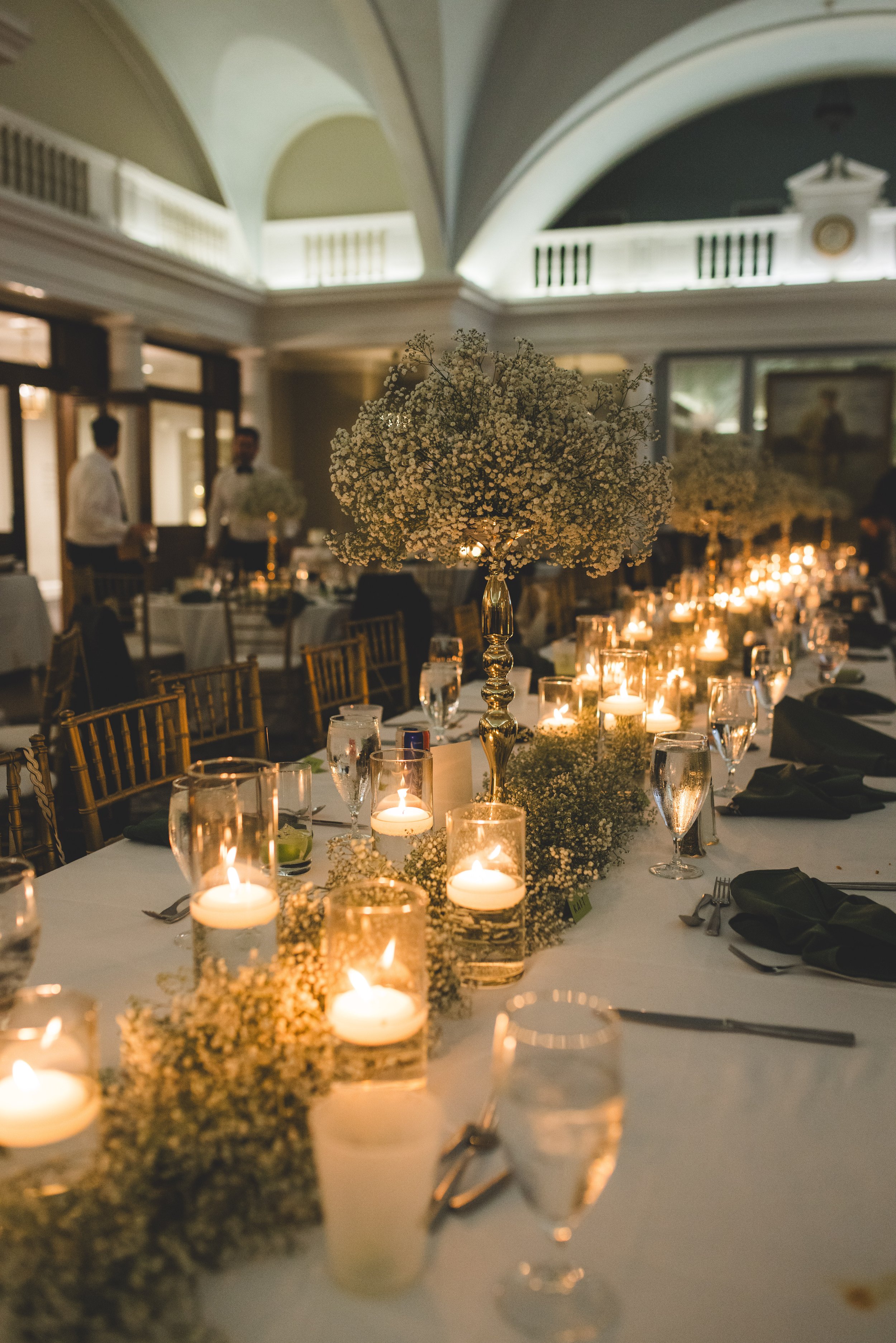 Elegant reception table decorated with white flowers, candles, and glassware in a dimly lit room with arched ceilings and framed paintings.