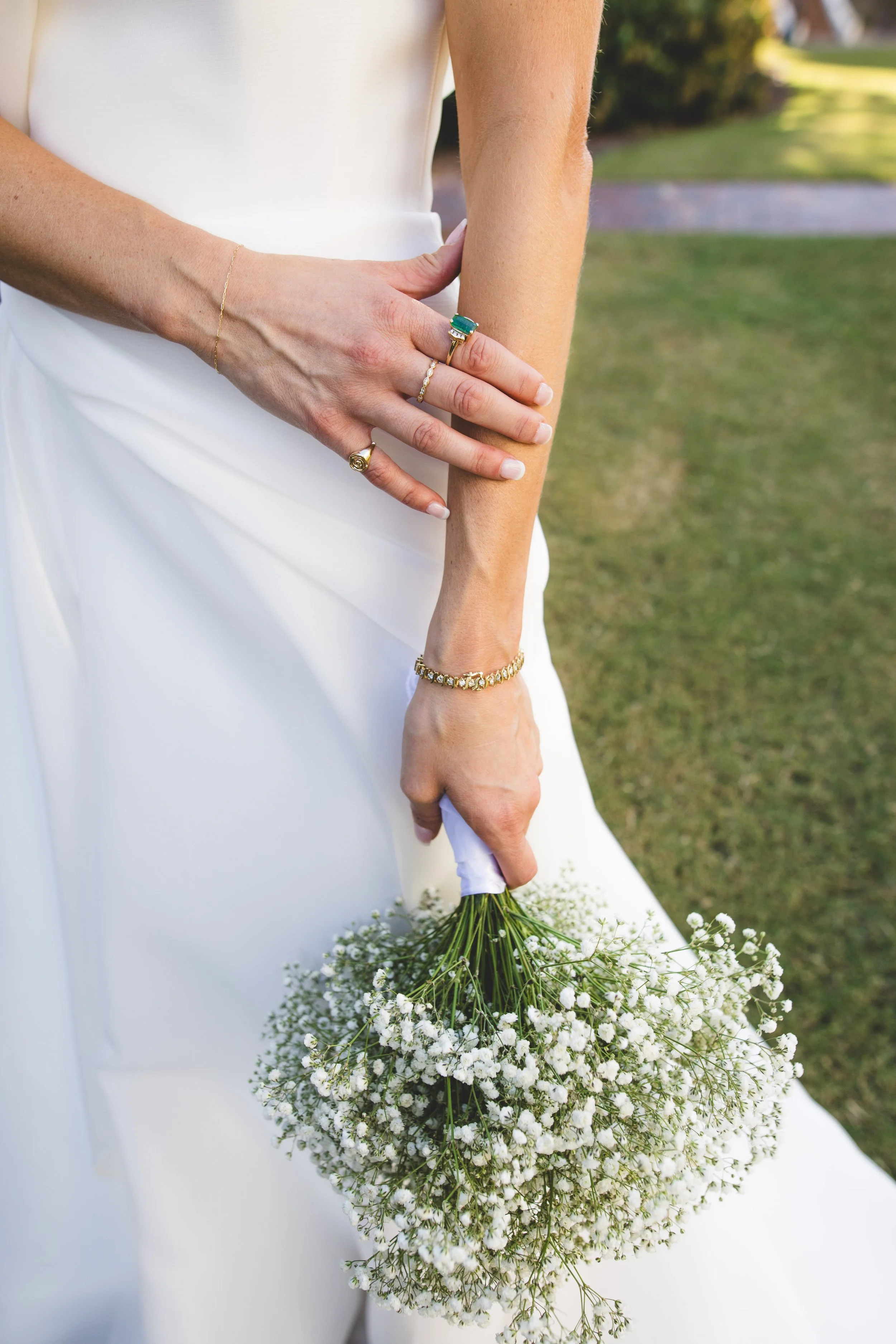 Close-up of a bride's hands holding a bouquet of white baby's breath flowers, with her wearing rings and bracelets, dressed in a white wedding gown outdoors.