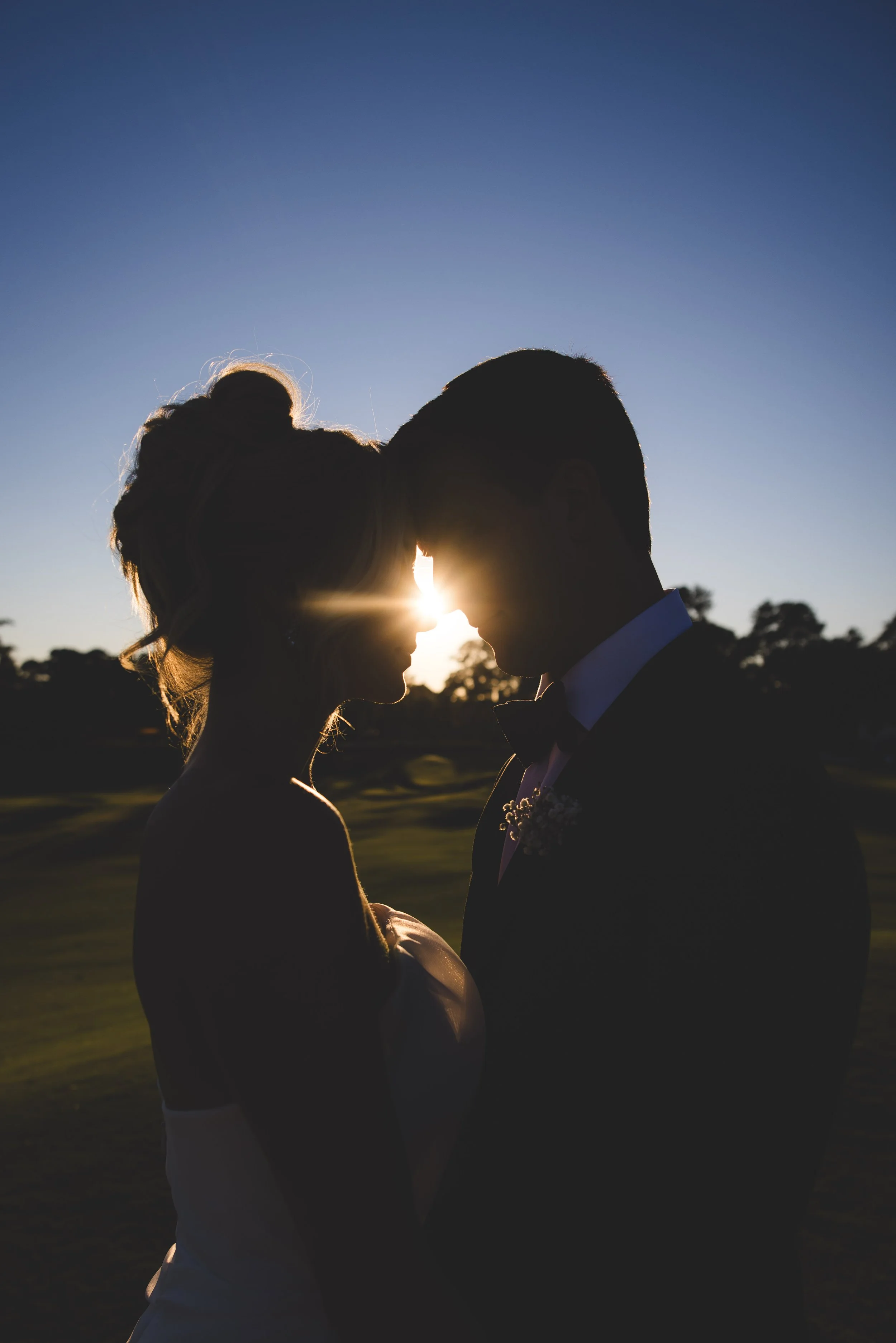 Silhouette of a bride and groom facing each other at sunset, with their foreheads touching, outdoors with trees in the background.