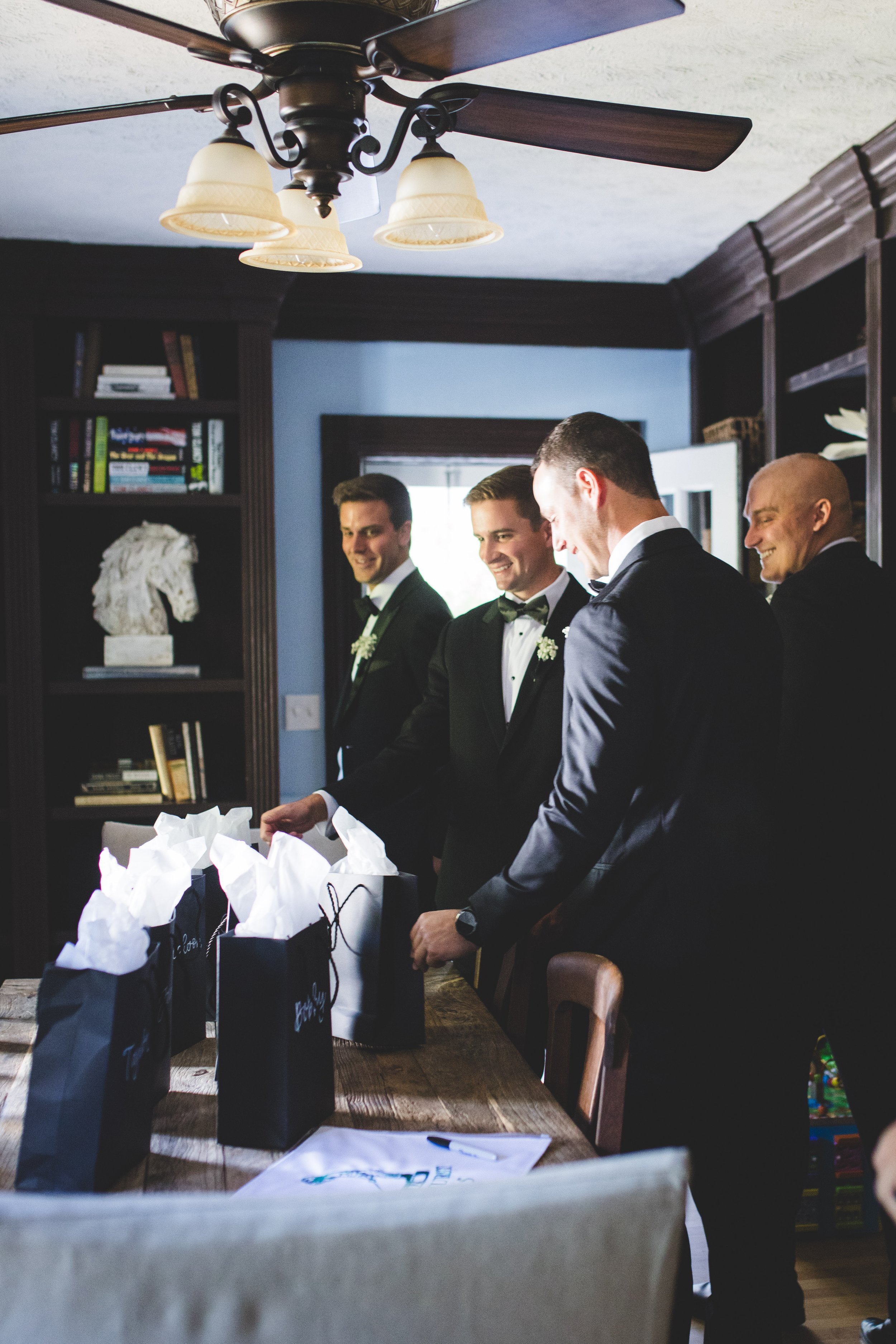 Four men in tuxedos gathered around a table with gift bags, inside a room with dark wood shelves and a ceiling fan.