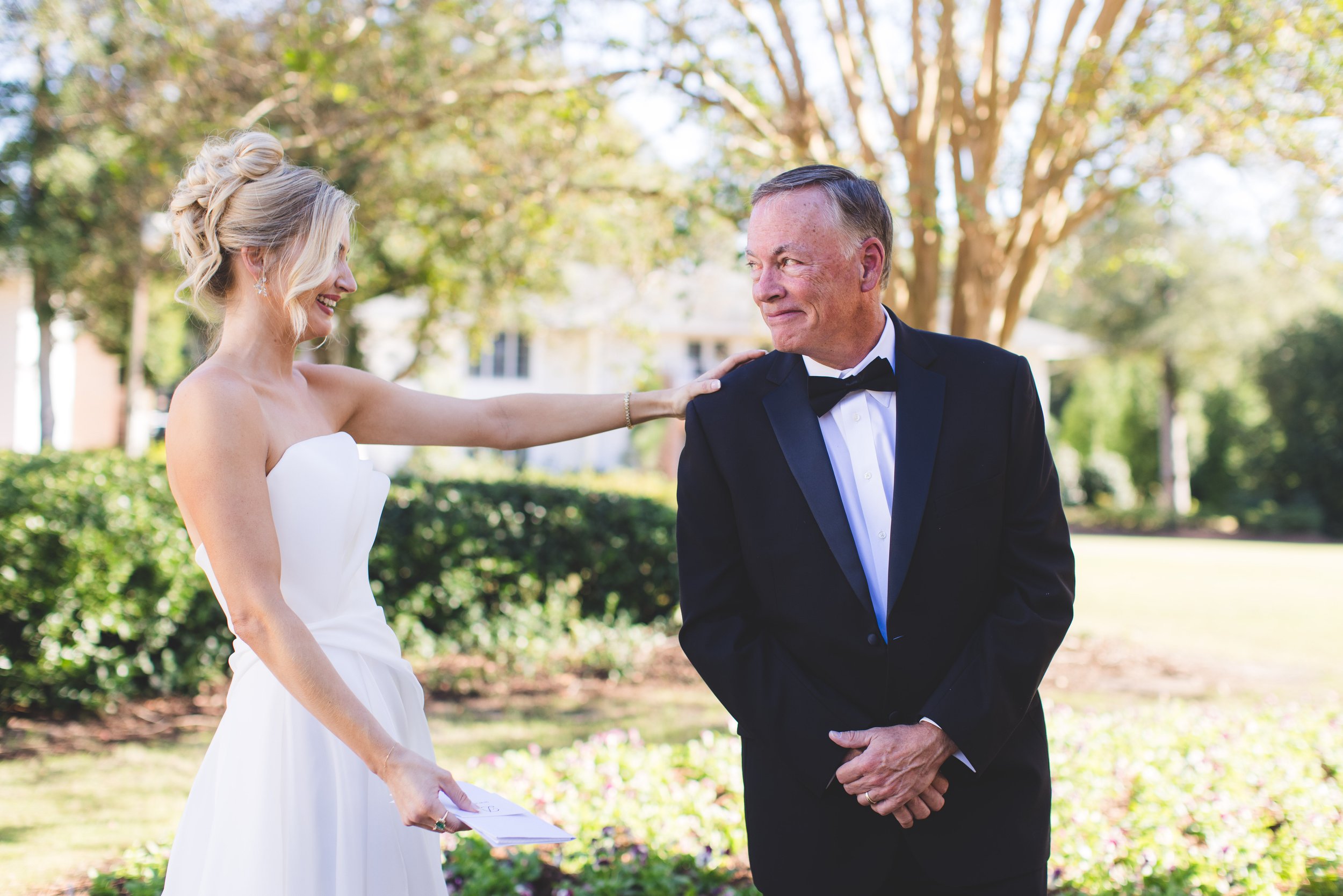 A bride and an older man, possibly her father, during a wedding ceremony outdoors. The bride is smiling and touching the man's shoulder, holding a piece of paper, while the man is smiling back with his hands clasped in front of him. Trees and greener