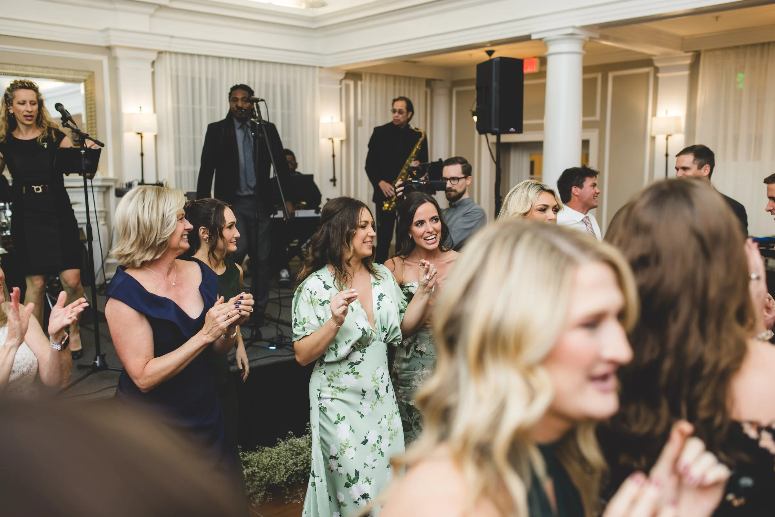 Guests dancing and clapping at a wedding reception with a live band performing in the background in an elegant venue.