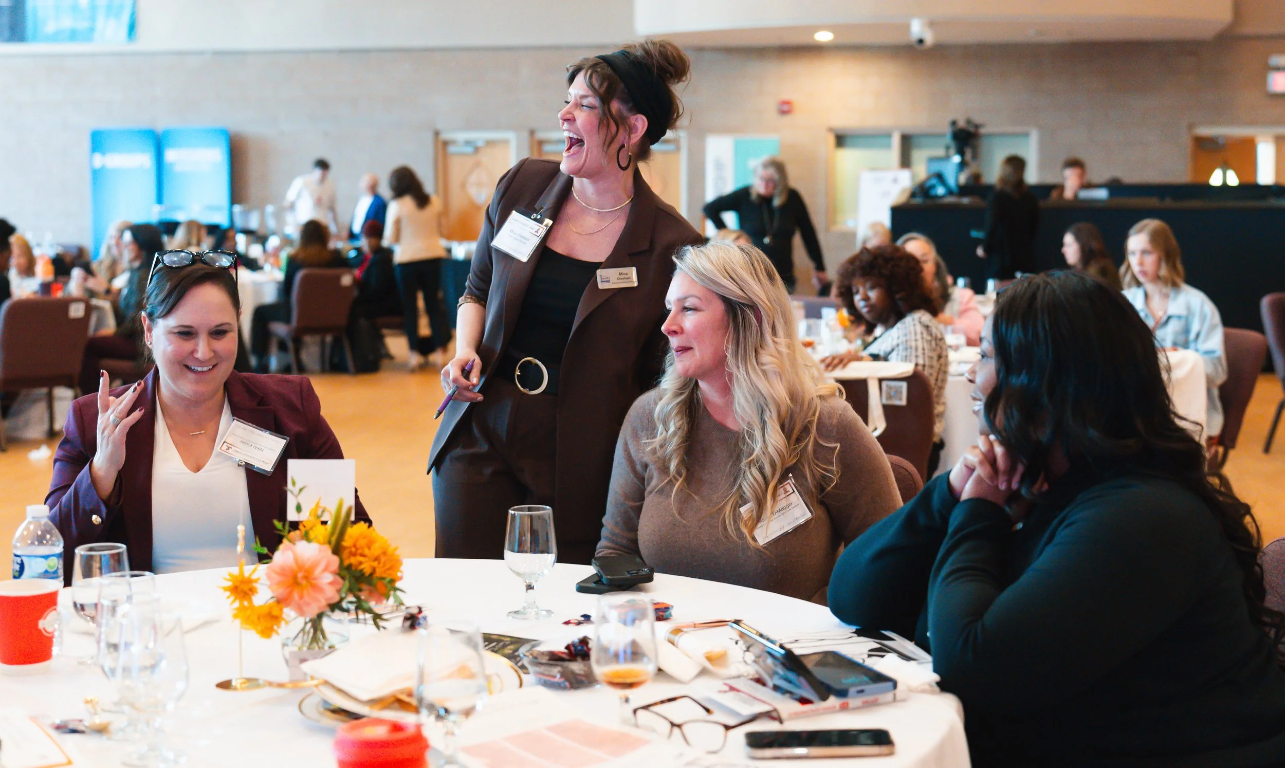 A woman with curly brown hair wearing a black headband and a brown blazer is laughing and talking with three women seated at a table during a conference or event, with other people and tables visible in the background.