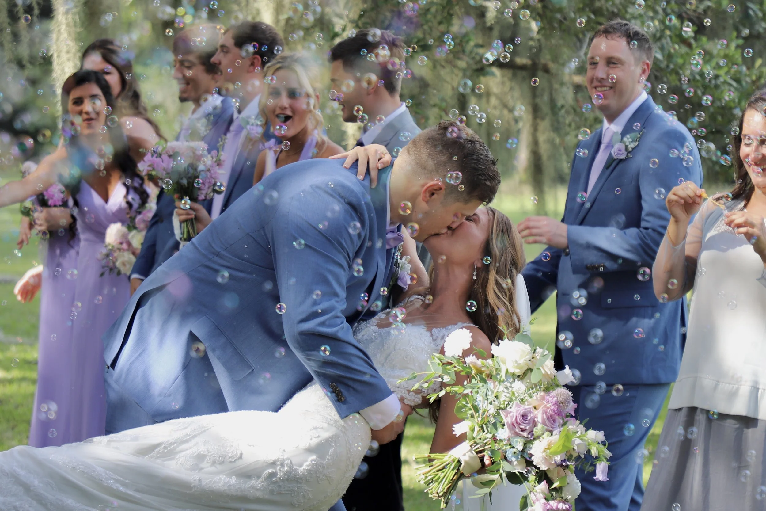 A newlywed couple shares a kiss at their outdoor wedding, surrounded by wedding party members and floating bubbles, with greenery in the background.