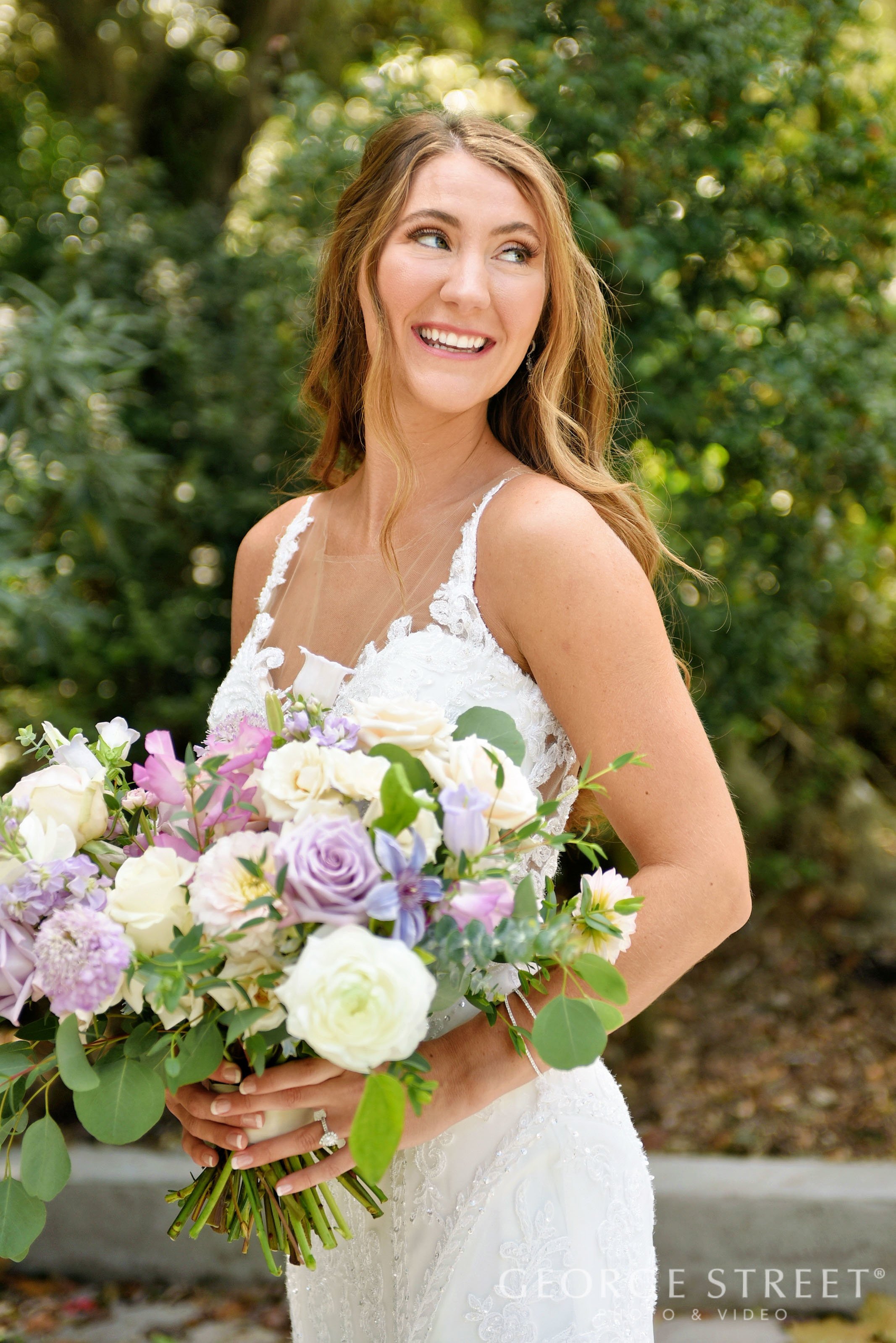A smiling bride with wavy, light brown hair holding a bouquet of white, purple, and pink flowers outside with green foliage in the background.