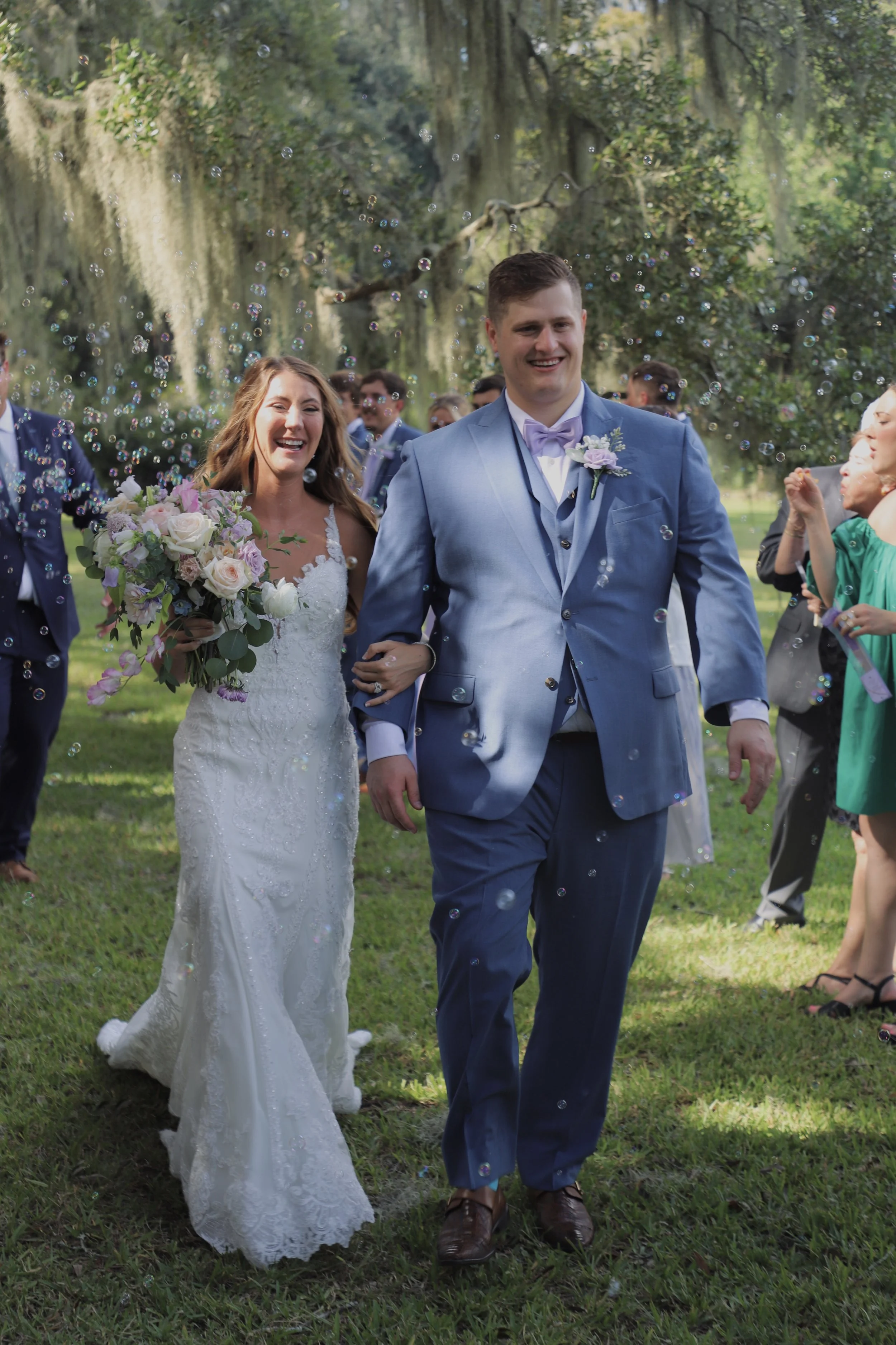 A newlywed couple walking outside through an outdoor wedding celebration, surrounded by guests. The bride is holding a bouquet of flowers, and the groom is dressed in a blue suit. They are smiling and happy, with bubbles floating in the air.