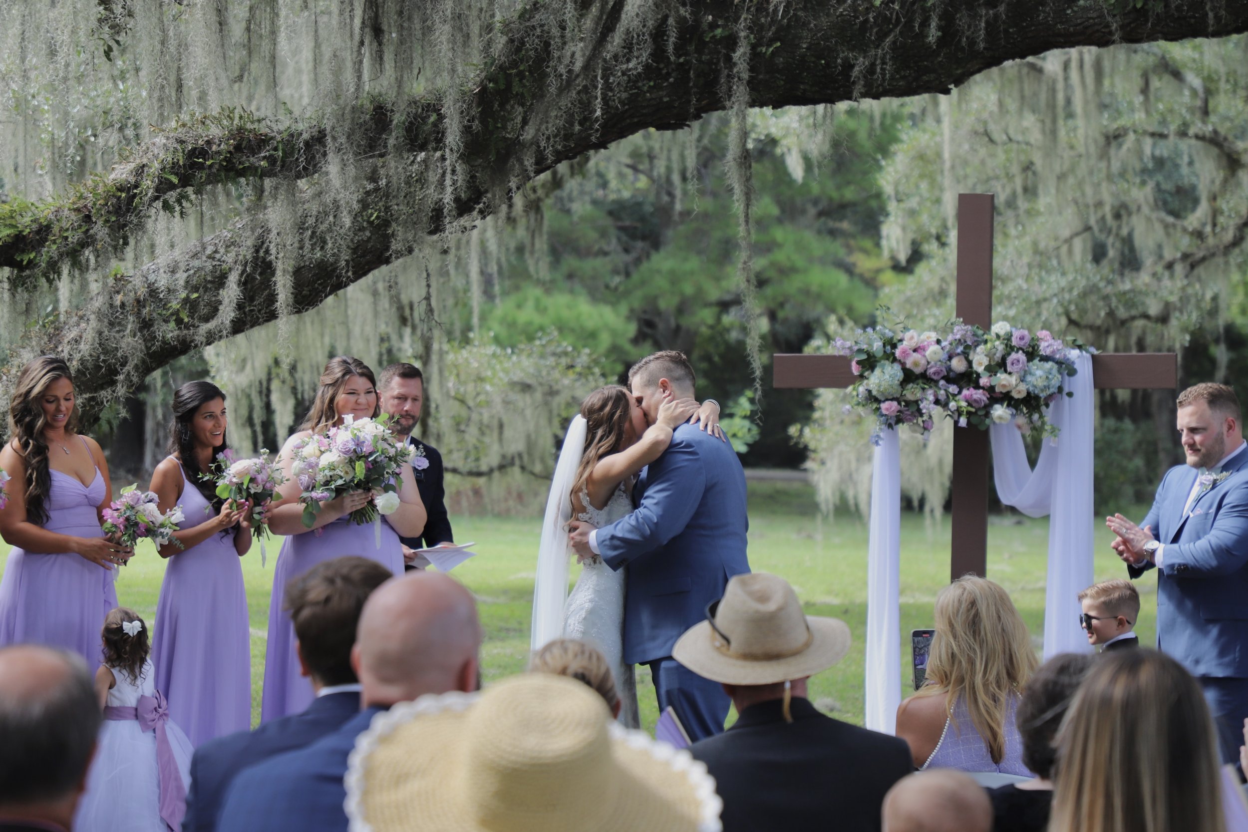 A wedding ceremony outdoors with the bride and groom kissing under a large tree with Spanish moss, surrounded by friends and family.