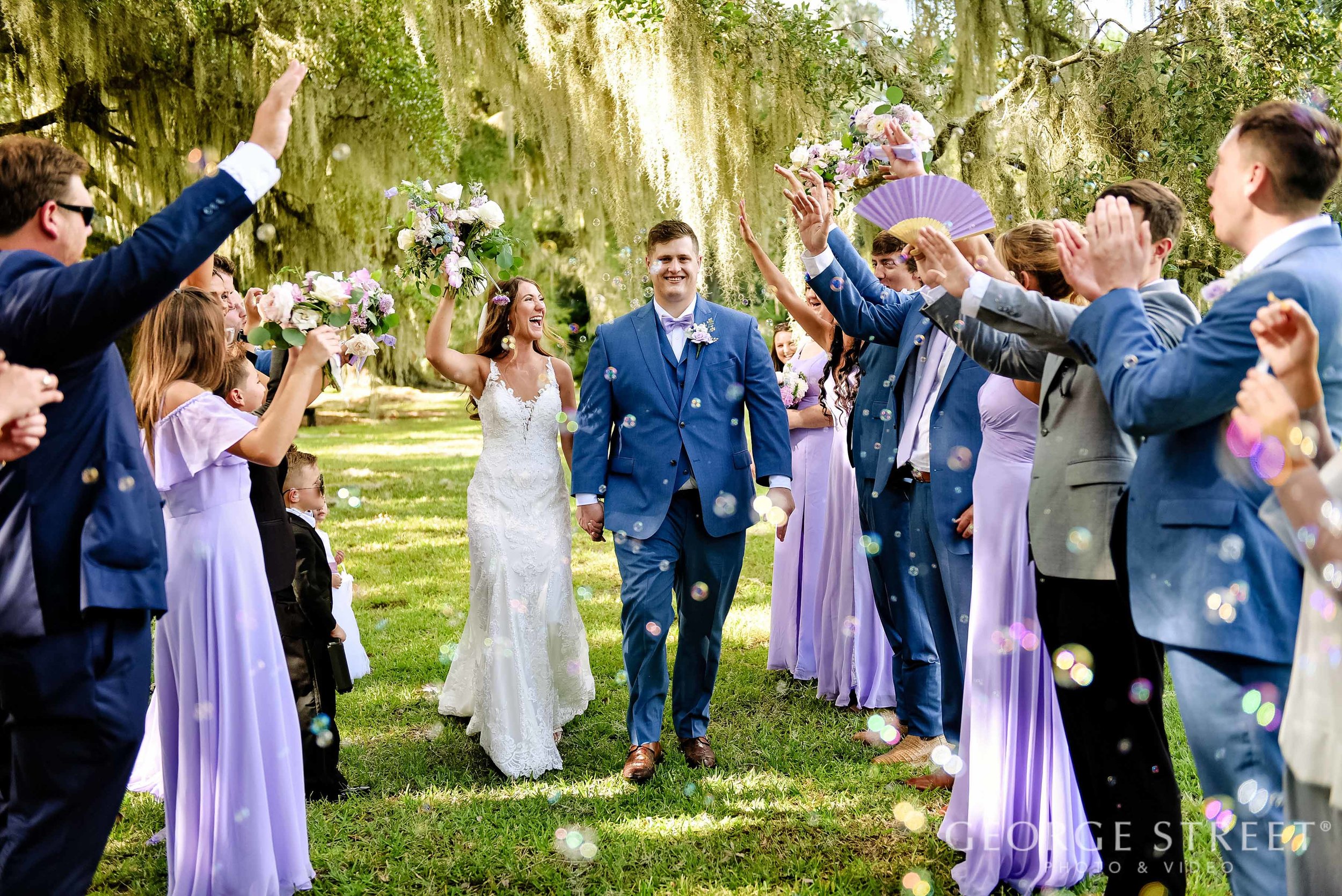 A bride and groom walking hand in hand down a grassy aisle surrounded by wedding guests in formal attire, celebrating with purple flowers and confetti on a sunny day in a park with hanging moss from trees.