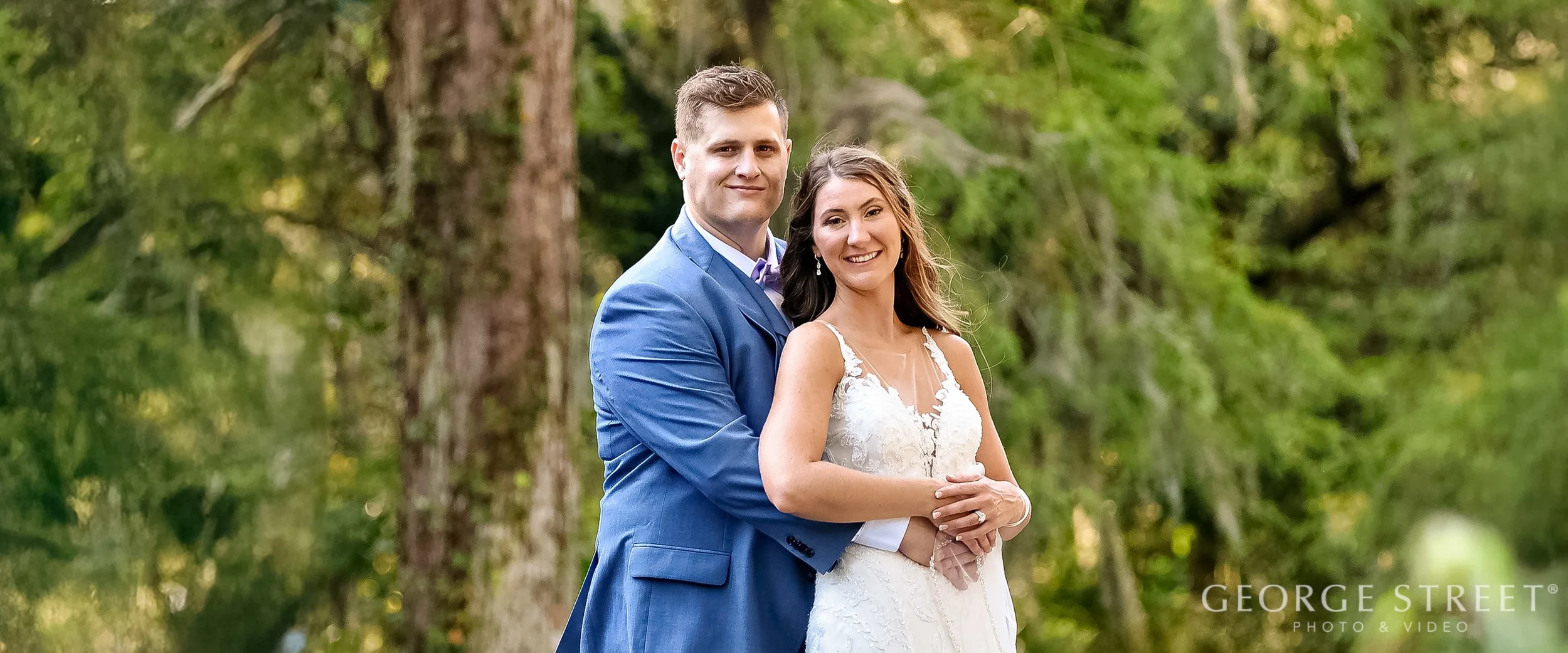 A happy bride and groom in wedding attire outdoors, standing close together amid green trees.