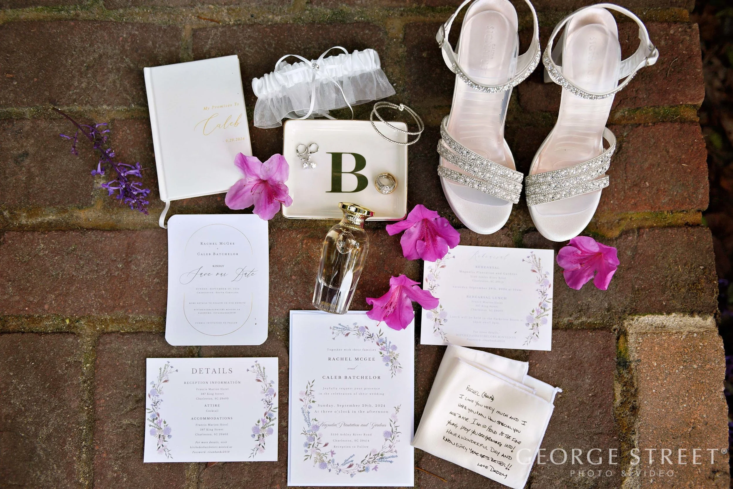 Wedding accessories including white shoes with rhinestones, jewelry, a perfume bottle, floral petals, and wedding invitation and details on a brick surface.